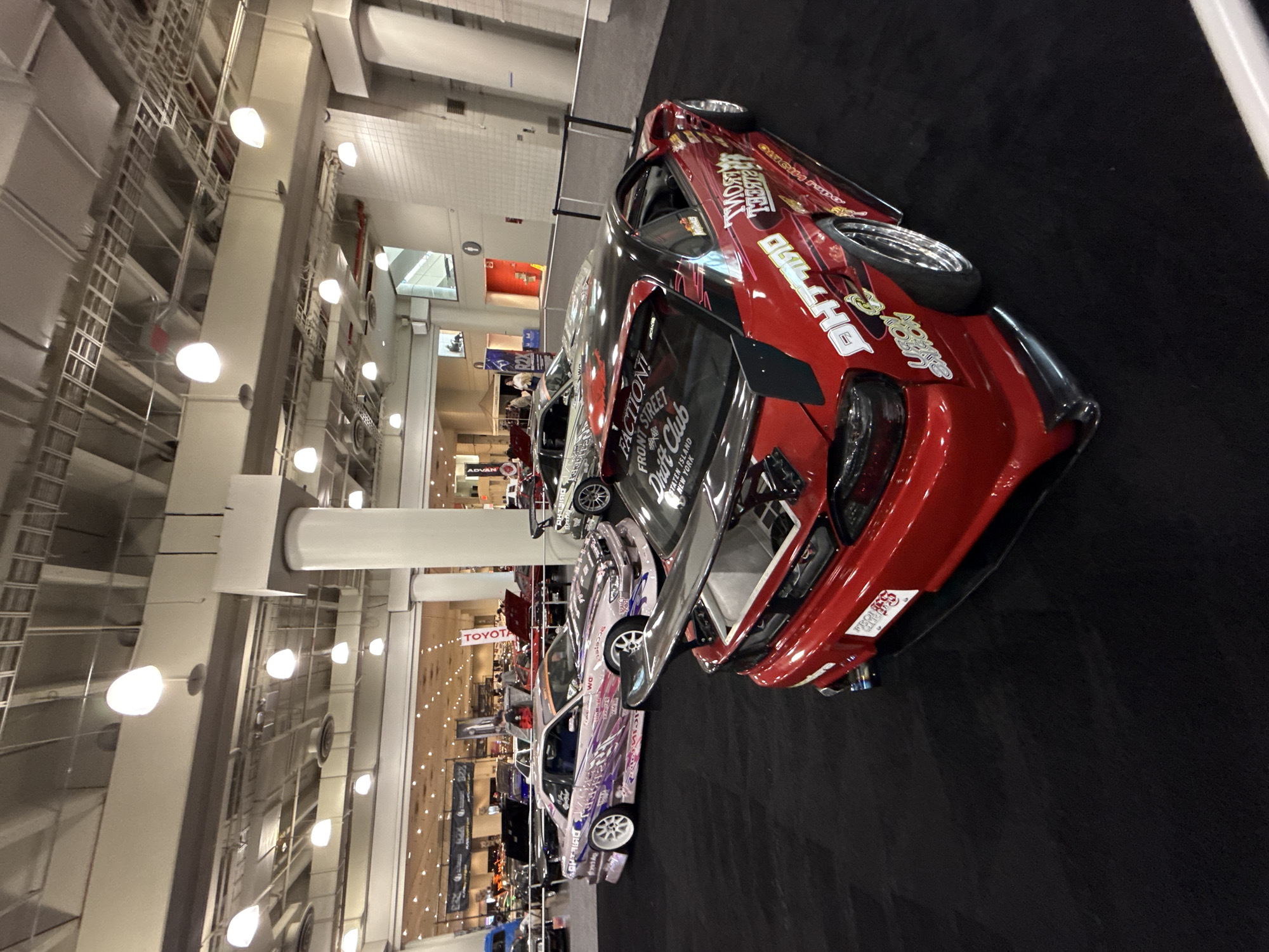 Here is a description of the image:
The image shows a line of customized cars, with a bright red car prominently featured in the foreground. This car has several sponsor decals on it, including "DH Tilt," and "Faction! Front Street Drift Club - Staten Island New York." Other cars, some with light purple coloration, are lined up behind it. The scene appears to be set inside a large exhibition hall or convention center, given the high ceilings, large pillars, and hanging lights. A "Toyota" sign is visible in the distance. Banners with "ADVAN" are visible on the left side of the image. The floor is covered in black carpet. The city and location are unknown.