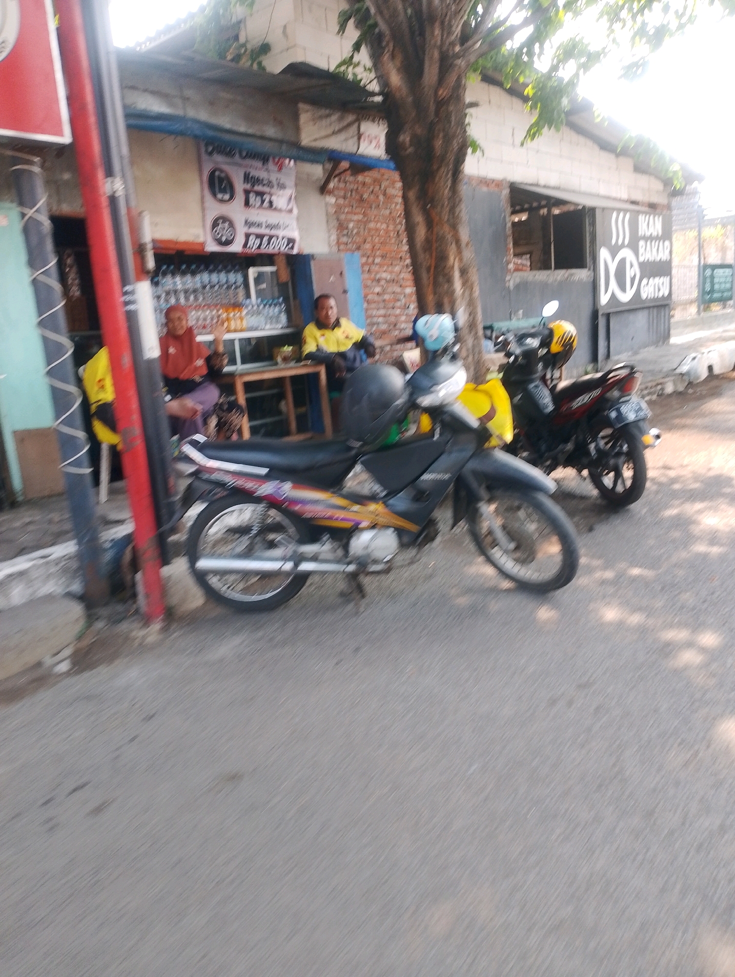 The image shows a street scene in front of small shops in Probolinggo, Indonesia. Two motorcycles are parked on the asphalt road. One motorcycle, in the foreground, is black with colorful decals on its side. The other motorcycle, behind it, is black with a red tail. In the background, a woman in a reddish-orange hijab and a person in a yellow shirt are sitting at a table outside one of the shops. Another person in a yellow shirt is visible standing. Signs are visible on the buildings, one appears to be advertising "Ikan Bakar Gatsu" (Grilled Fish Gatsu), and another seems to be an advertisement with prices and icons. The buildings have simple brick and concrete construction, with corrugated metal roofs. A tree with green leaves is to the left of the "Ikan Bakar Gatsu" sign, casting shadows on the ground. The overall environment suggests a commercial area with a relaxed, possibly local atmosphere.