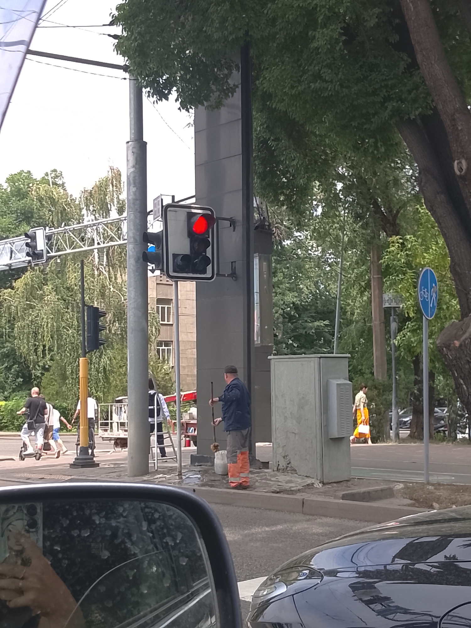 The image depicts a street scene in Almaty, Kazakhstan, focusing on a pedestrian crossing with traffic signals. A traffic light is prominently displayed, showing a red signal. To the left, a person on an electric scooter is crossing the street, accompanied by another pedestrian. Further down the street, another traffic light is visible. A worker in orange pants and a dark jacket is standing near a utility box, holding a broom. A woman in an orange outfit is walking in the background. A bicycle lane sign is also present. The scene is framed by trees and urban architecture. The foreground shows the reflection in a car's side mirror, revealing a person's hand with rings.