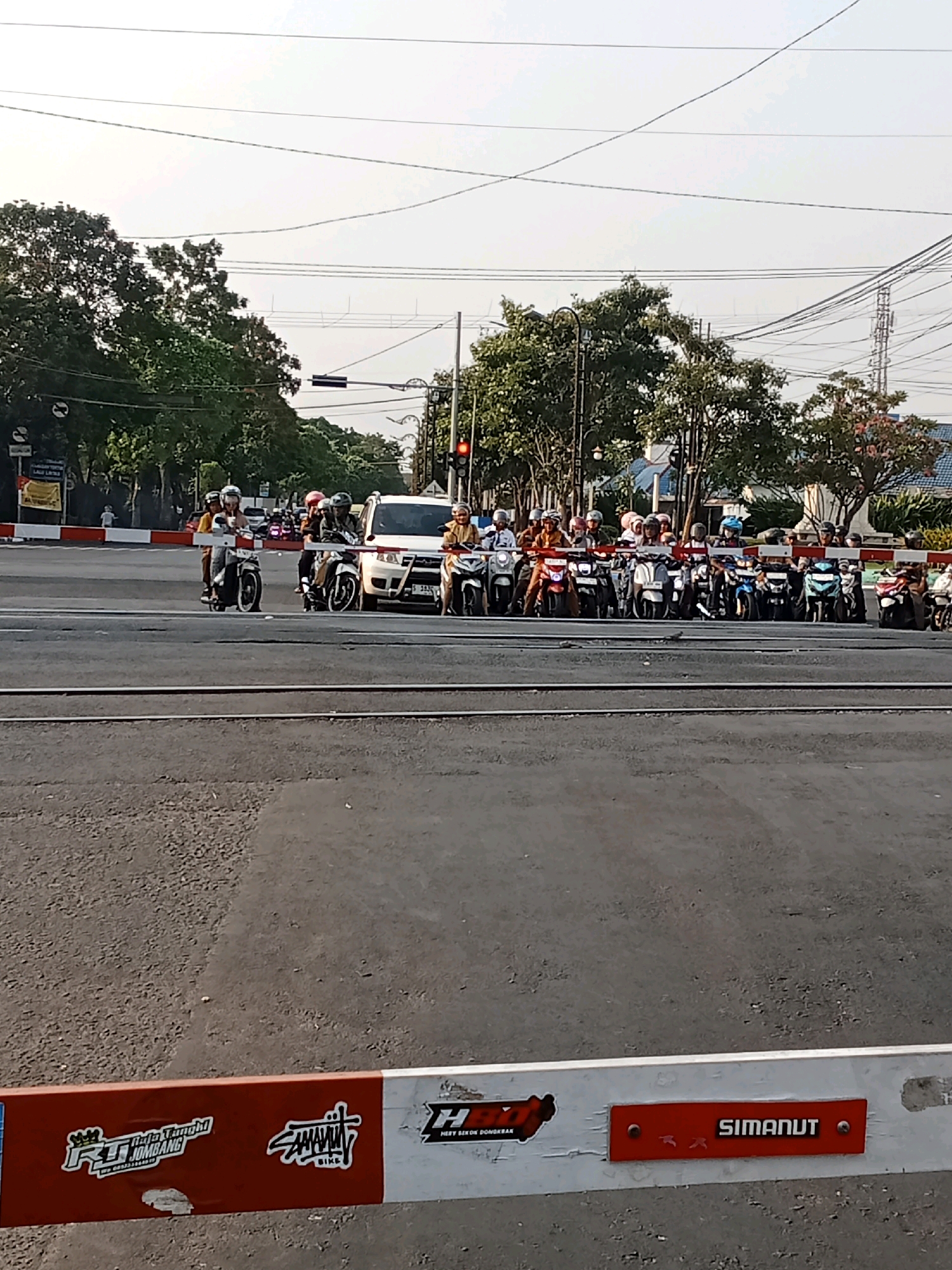 At a railway crossing in Kaliwungu, Indonesia, a group of motorcyclists waits for a train. The motorcyclists, wearing helmets, are stopped behind a red and white barrier. The barrier appears to be a railway crossing gate. In the background, there are trees, a white van, and traffic lights. The sky is clear and the scene is illuminated by sunlight.