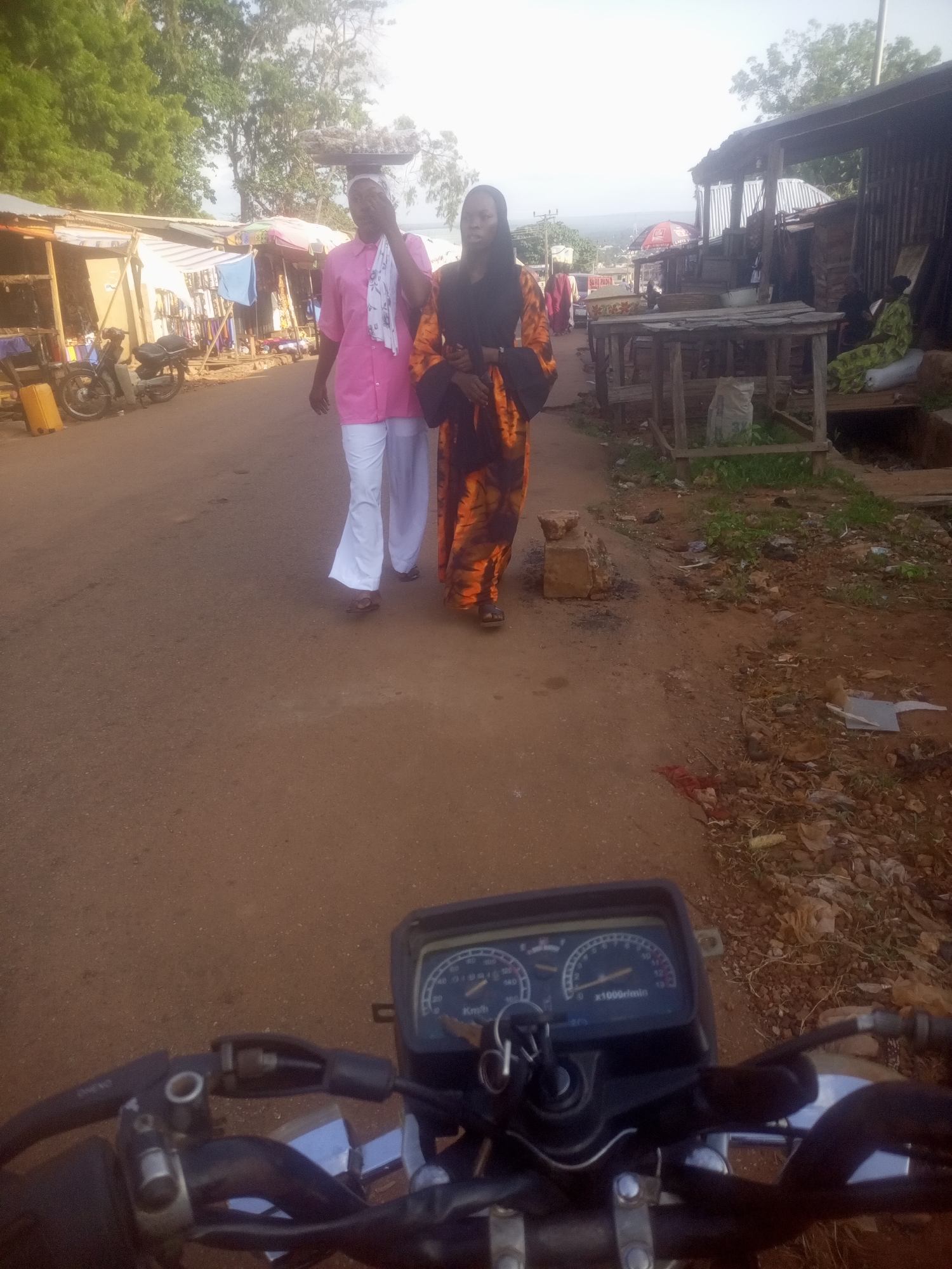 This image depicts a street scene in Ankpa, Nigeria. In the foreground, the handlebars and dashboard of a motorcycle are visible, suggesting the photo was taken from the perspective of a rider. The road is unpaved and dusty, characteristic of many streets in the region.
On the right side of the road, there are market stalls with wooden tables and corrugated metal roofing. Some stalls appear to be selling goods, with various items displayed. One stall has a person in a green garment sitting behind a table, and sacks of materials are visible. The ground around the stalls is littered with debris, including plastic bags, leaves, and other waste.
On the left side of the road, more market stalls are set up, with tarpaulin coverings and displays of goods, including clothing and a yellow fuel container. A motorcycle is parked in front of one of these stalls.
Two women are walking away from the camera on the paved portion of the road. The woman on the left is wearing a bright pink top and white trousers, carrying a tray of items on her head. She is also holding a white cloth. The woman on the right is wearing an orange and black patterned dress and a dark headscarf. They appear to be walking towards the market area. The general atmosphere suggests an active marketplace, possibly in the afternoon given the lighting.