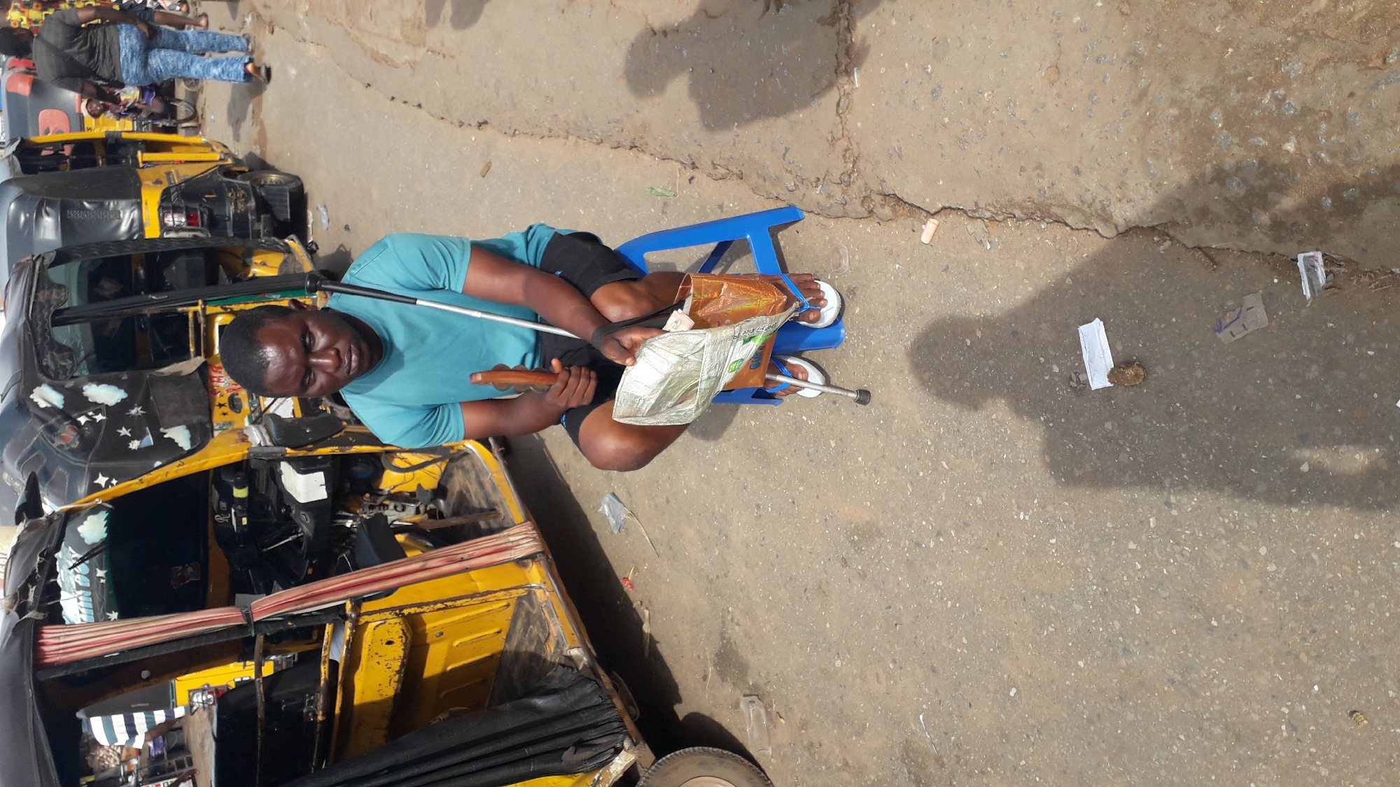 In Nkpor, Nigeria, a Black man with a dark complexion is seated on a blue plastic stool next to a yellow auto-rickshaw. He is wearing a teal t-shirt and black shorts, with white flip-flops on his feet. He holds a white cane in his right hand, which rests on the ground, and a brown object, possibly a wooden staff, in his left hand. In front of him, a crumpled plastic bag filled with folded papers, likely money, rests on his lap. The scene appears to be a street, with a rough, paved ground and the auto-rickshaw's tires visible. Other auto-rickshaws and people are present in the background, indicating a bustling urban environment. The sunlight casts a shadow of the photographer on the ground.