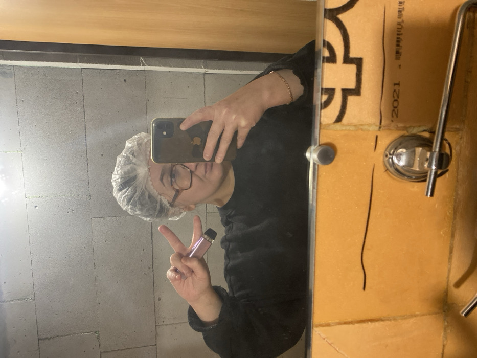 An individual is captured in a mirror selfie, reflecting their upper torso and head within an indoor setting. The person wears a clear, disposable shower cap, large rectangular brown-framed glasses, and a dark long-sleeved top. Their left hand is raised, forming a peace sign, and holds a pink cylindrical object with a black tip, possibly a vape pen or cosmetic tube. A thin, beaded bracelet adorns their right wrist. The individual's right hand holds a dark-cased iPhone, obscuring part of their face while taking the photograph. The wall reflected above the mirror consists of light grey, rectangular masonry blocks. To the right, a light brown wooden surface or door frame is visible. Below the mirror, a surface is covered with a textured, light orange material, possibly insulation or paneling, exhibiting black markings including a wavy line and partially visible text "2021." Chrome fixtures, including a faucet and a towel bar, are mounted on this orange surface. The specific location within the city of Chaikovsky, Russia, is not discernible from the image.