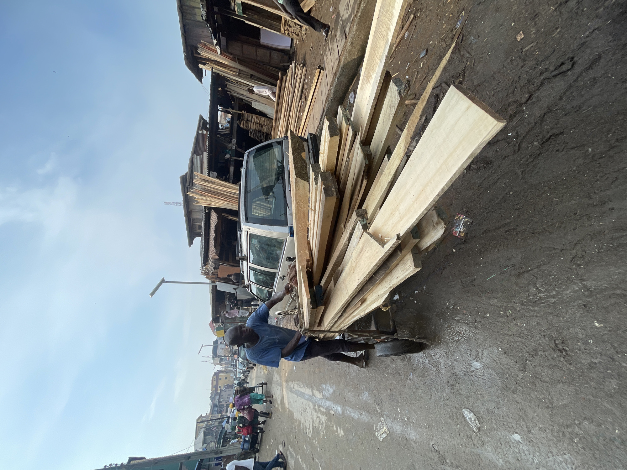 In Lagos, Nigeria, a man is securing a large stack of wooden planks onto the roof rack of a silver van. He is wearing a short-sleeved blue shirt and dark pants. The street is wet and muddy. Other people are visible in the background, along with shops or stalls constructed from wood. The sky is overcast.