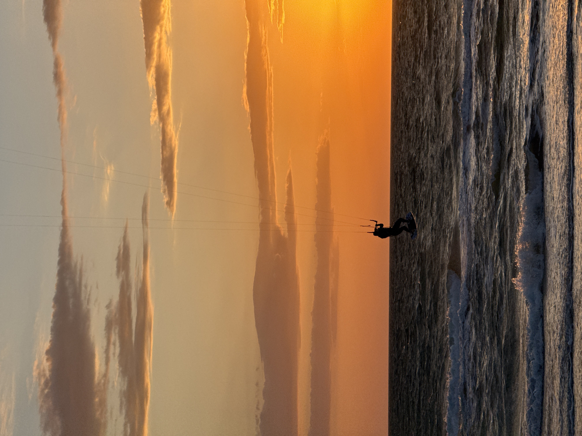 A kiteboarder is in the air on the ocean with a bright orange sunset. The sky is mostly covered in a dark orange glow from the setting sun. The top right section of the image is the ocean which is dark and filled with waves. A few wispy clouds are scattered in the sky. The location is Unknown, Unknown.