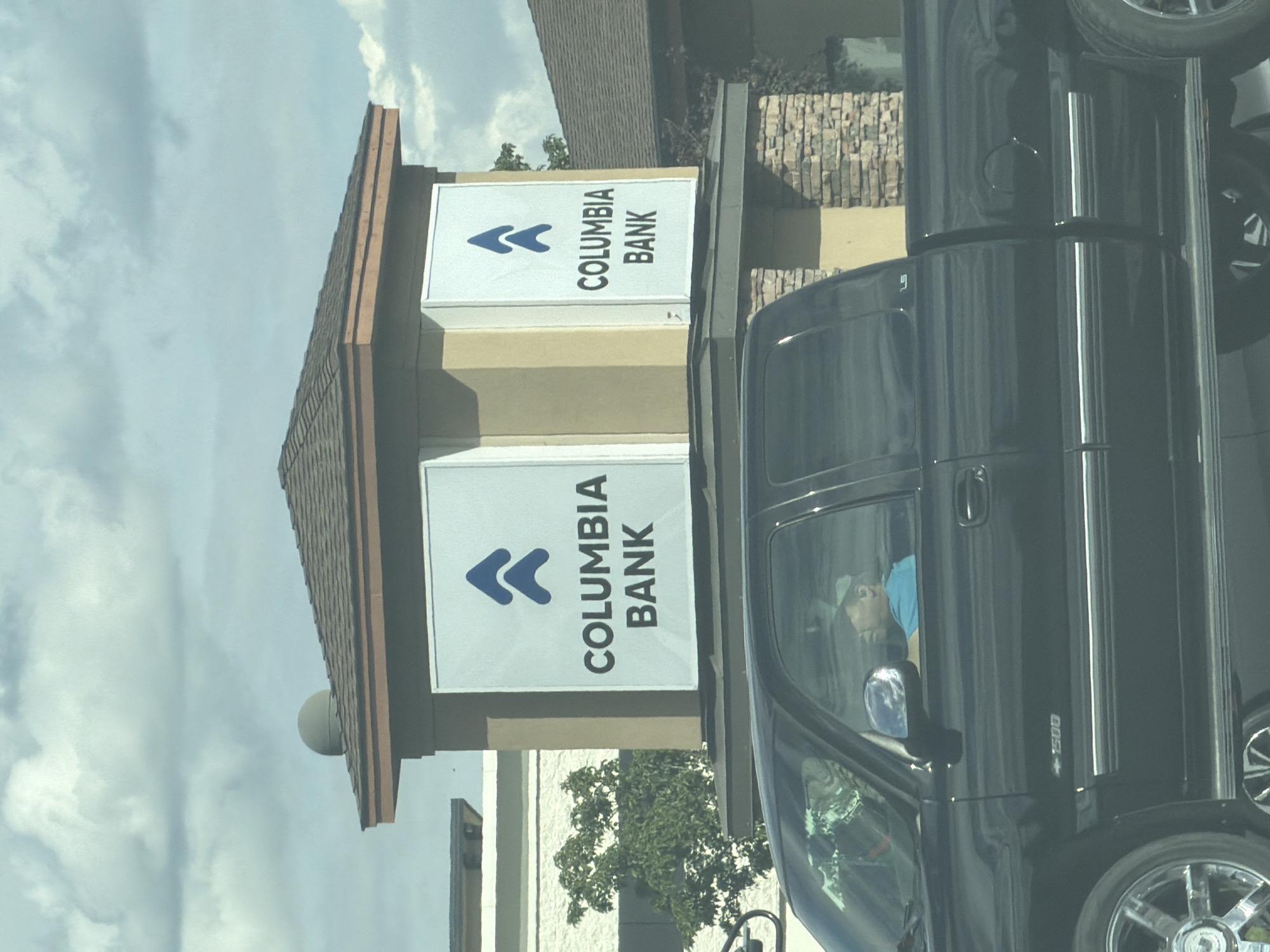 A black pickup truck is parked in front of a Columbia Bank building. The truck has tinted windows, chrome wheels, and "1500" emblem on the side. A person wearing a blue shirt and a baseball cap is visible in the driver's seat. The bank building is light tan with a brown roof and has two signs with the Columbia Bank logo and name. The background shows a cloudy sky and trees. The scene is located in Pasco, United States of America.
