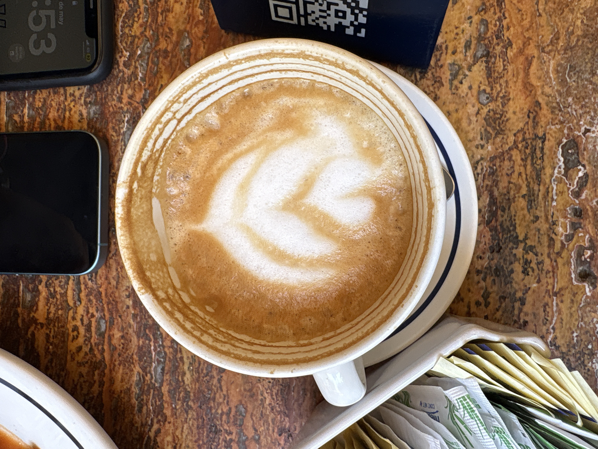 A top-down shot captures a latte with latte art, nestled on a saucer. Adjacent to the latte is a container filled with sugar packets, a partially visible phone, and a dark blue object with a QR code. The surface is wood and appears to be a table. The photo may have been taken in Mexico City, Mexico. The time on the phone reads 12:53.