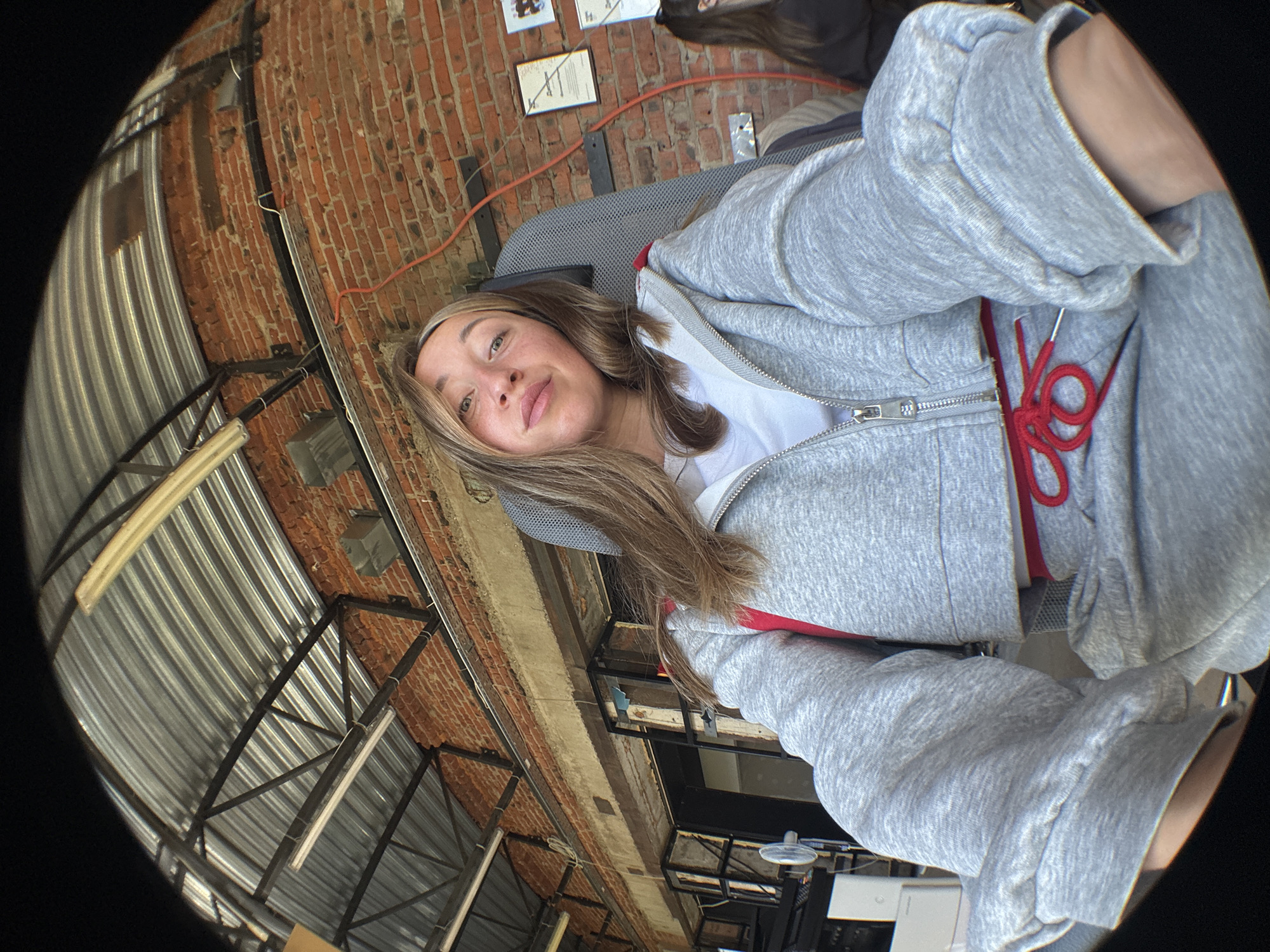 A young woman with long, light brown hair is seated in a chair indoors, facing the camera. She is wearing a grey cropped zip-up hoodie over a white t-shirt and grey sweatpants with a red drawstring. The background consists of a brick wall, a corrugated metal ceiling with exposed beams and fluorescent lights, and some framed documents on the wall. A red cable is visible running along the brick wall. The image appears to be taken with a fisheye lens, causing distortion. The scene suggests an indoor industrial or workshop setting, possibly a studio or garage. No specific location in Chaikovsky, Russia is discernible from the image itself.