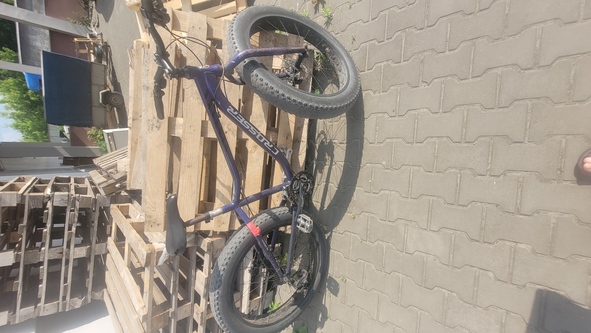 A purple bicycle is leaning against a stack of wooden pallets outdoors. The bicycle has fat tires and appears to be a mountain bike. The brand name "CRUSSER" is visible on the bicycle frame. The scene is set on a paved area with interlocking bricks, suggesting an outdoor setting like a yard or a street. The lighting suggests it is daytime, and the shadows cast by the bicycle and pallets indicate sunlight. There are no people prominently visible in the frame, although a foot wearing a sandal is partially visible in the bottom right corner of the image. The background shows more pallets and some greenery, hinting at a storage area or a place where materials are handled. The overall mood is casual and possibly depicts a moment of rest or storage.
