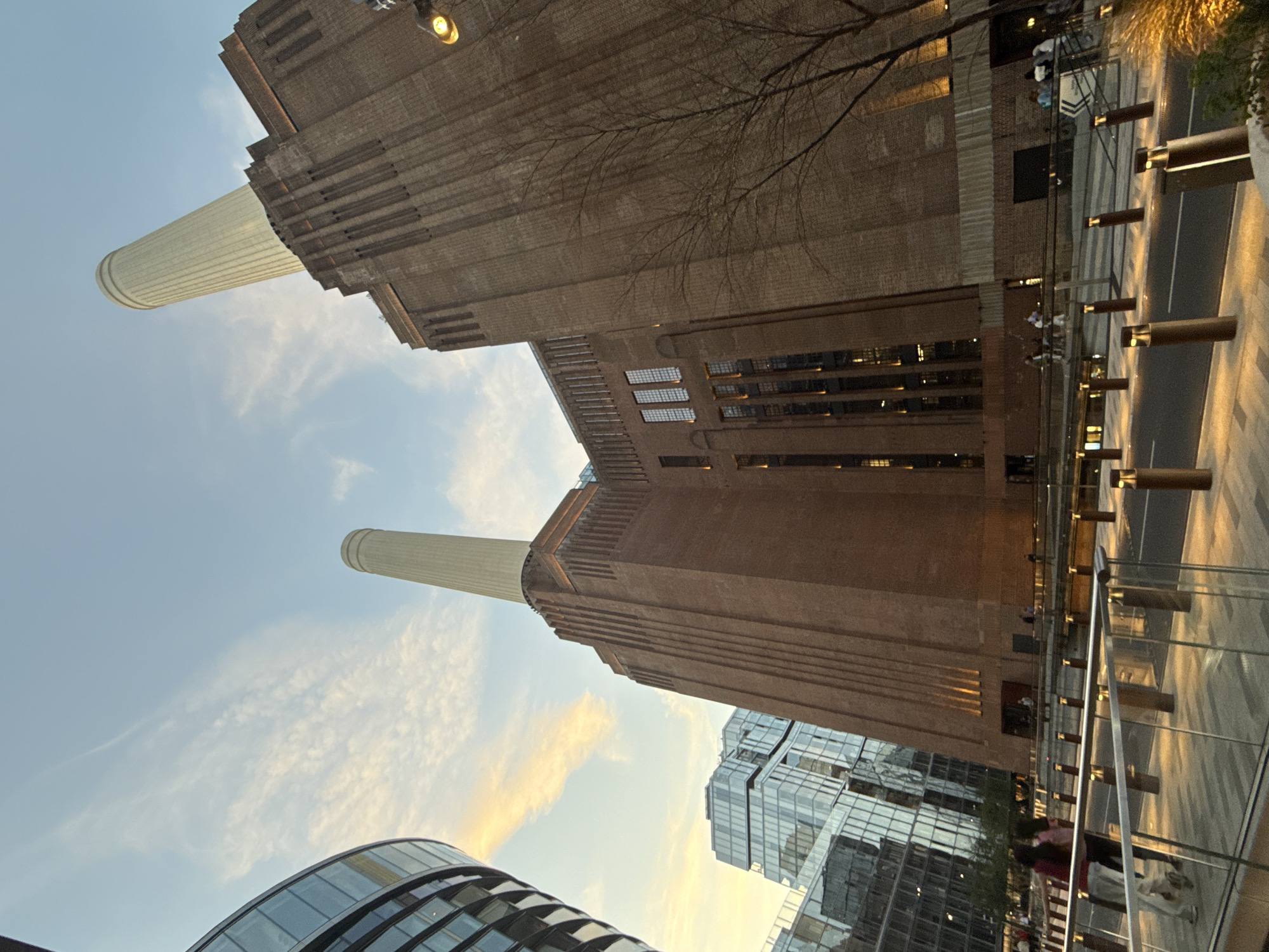 The image depicts the Battersea Power Station in London, United Kingdom, viewed from a low angle. The power station's brick facade and two of its chimneys are prominent against a blue sky with wispy clouds. Adjacent to the power station are modern glass-clad buildings. A pedestrian area with glass railings and bollard lighting is visible in the foreground, with people walking along the path.