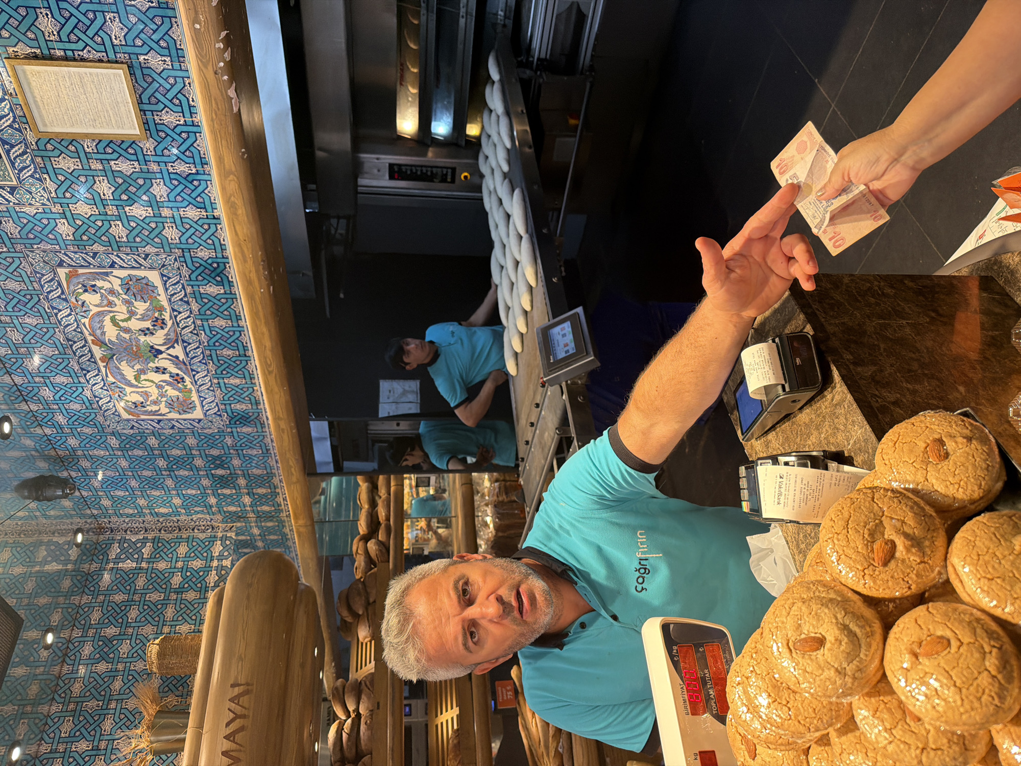 In a bakery in Kadıköy, Turkey, a male employee in a turquoise polo shirt with "çağrınfin" written on it, stands behind a counter. He has gray hair and a beard and appears to be speaking. In front of him are several stacked cookies wrapped in plastic, topped with almonds. To his right, a customer's hand is reaching out to give him a 10 Turkish Lira banknote. A credit card machine is visible on the counter. In the background, other employees are working, and loaves of bread are displayed on shelves. The walls are decorated with intricate blue and white tiles.