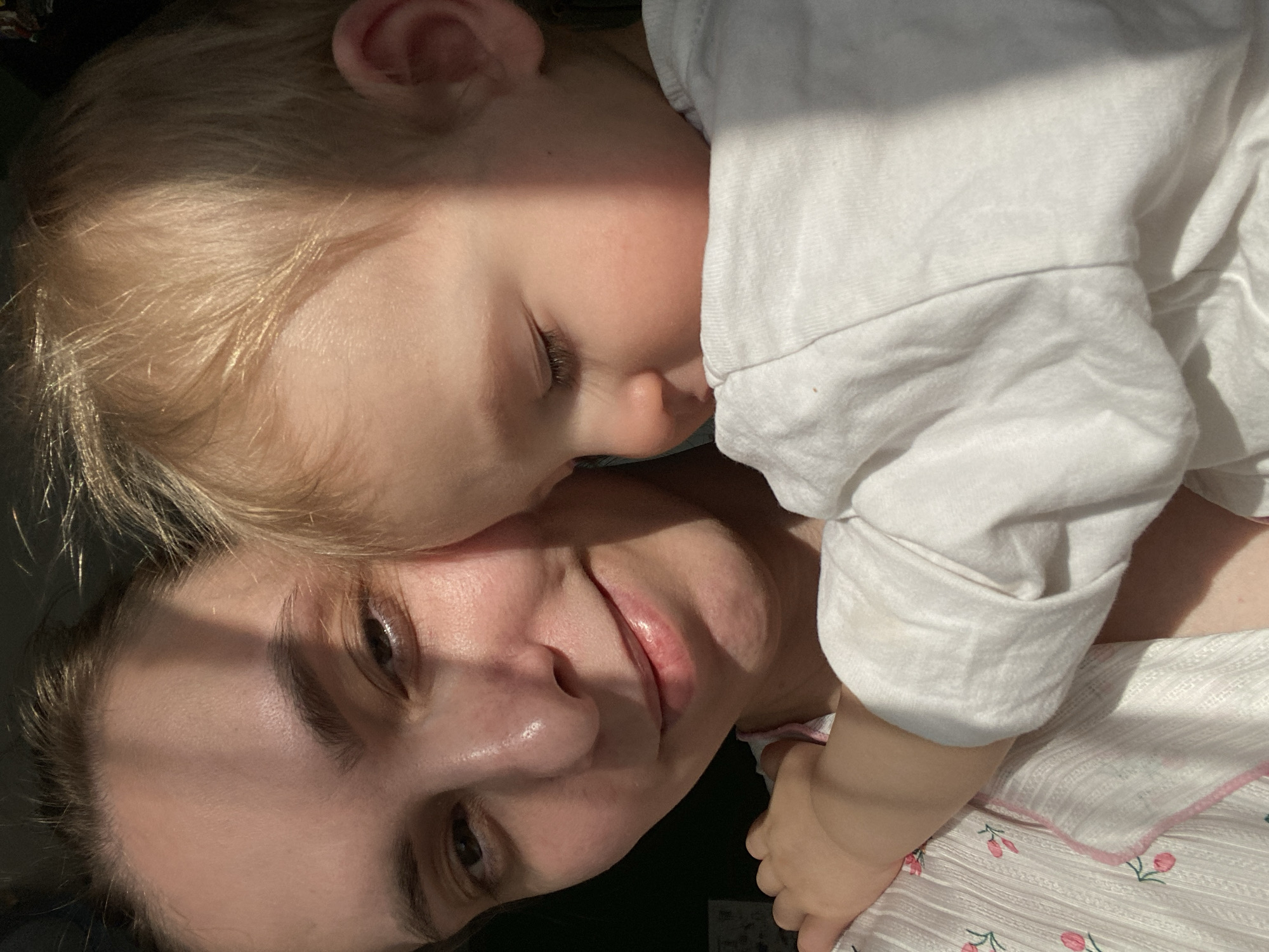 A woman is lying down with a young child resting on top of her. The child has fair hair and is wearing a white shirt. The woman has dark hair and is wearing a light-colored top with a floral pattern. The scene is brightly lit. The location is unknown and there is no indication it is Chaikovsky, Russia.