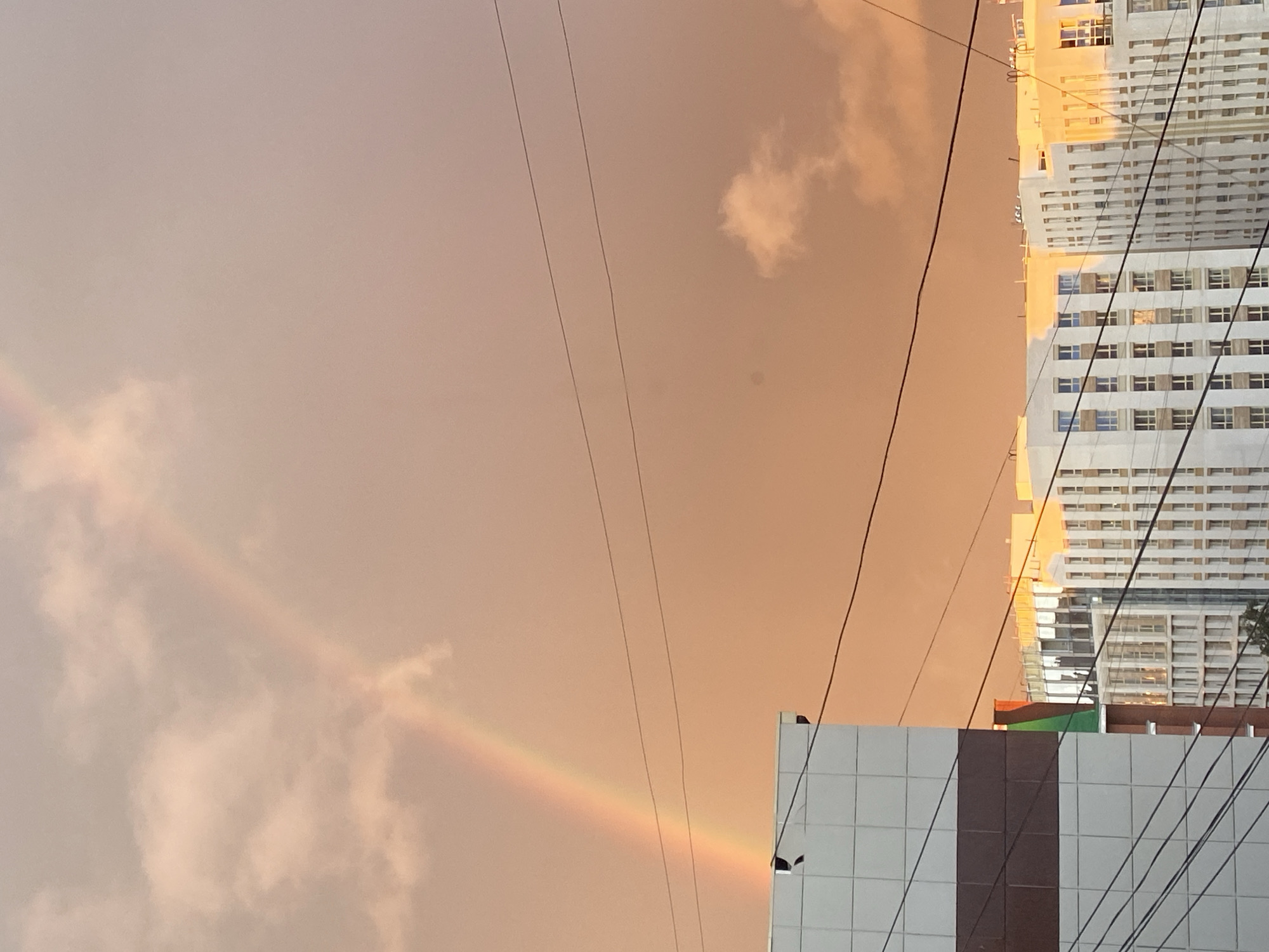 The image captures a view of the city of Perm, Russia, dominated by a pale orange sky with a visible rainbow arching across it. In the foreground, the corner of a modern building with a white tiled facade and a brown horizontal stripe is prominent on the left. Several black utility wires crisscross the frame, obscuring parts of the background. Beyond the foreground building, a row of contemporary apartment buildings with white facades and numerous windows stretches across the mid-ground and background. The setting appears to be a cityscape during daylight, possibly after a rain shower, given the presence of the rainbow. No people are visible in the image.