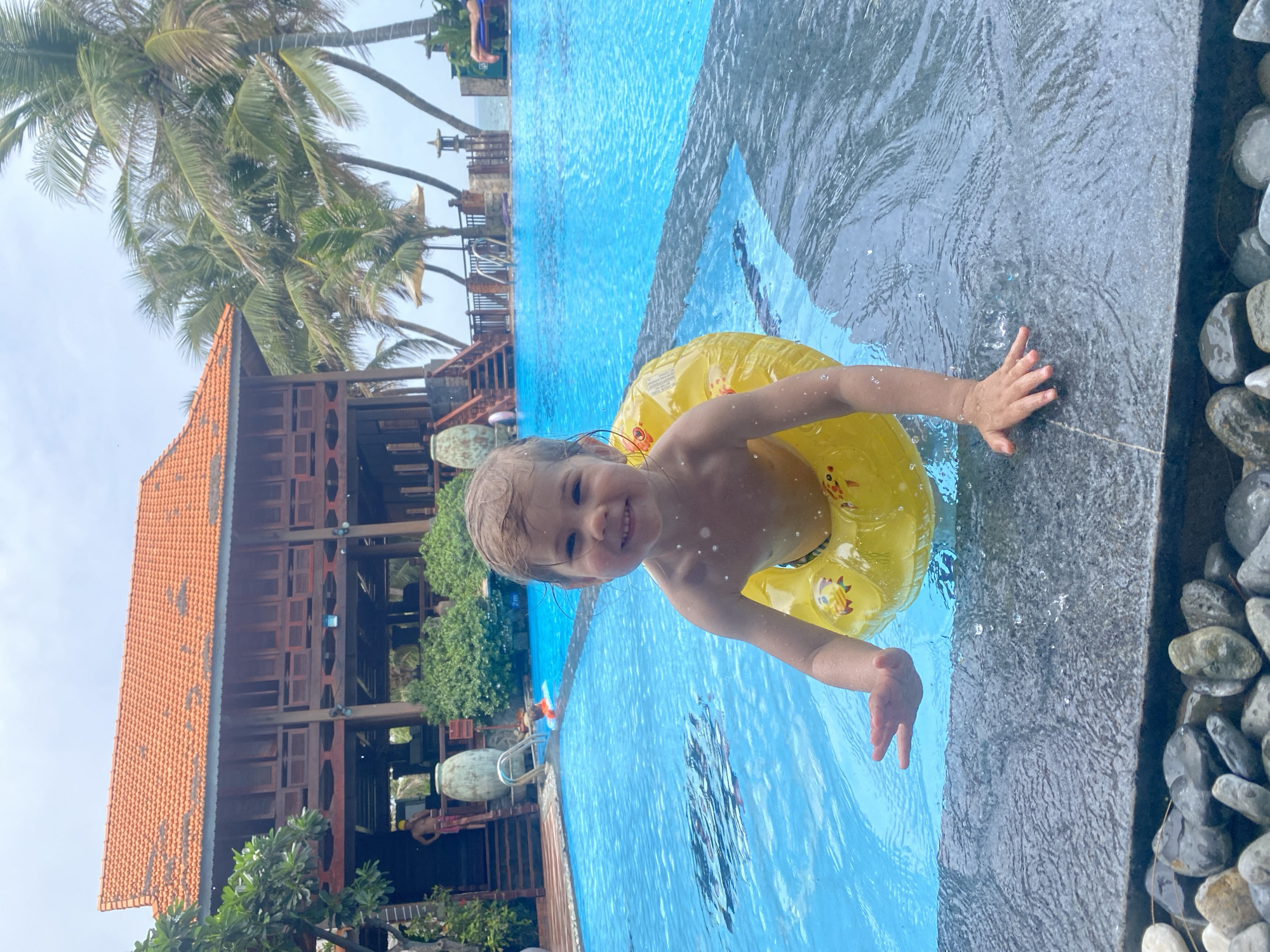 A young child with light hair is smiling directly at the camera, partially submerged in a swimming pool and wearing a vibrant yellow inflatable swimming ring. Water droplets are visible on the child's face and shoulders, and one hand rests on the dark, wet edge of the pool. The swimming ring has faint cartoon-like patterns.
The scene is an outdoor swimming pool, likely at a resort or hotel in Phu Thuy Ward, Vietnam. The pool water is clear and blue with visible ripples. In the background, to the left, stands a multi-story building with a distinctive reddish-orange tiled roof and wooden architectural details, hinting at a tropical or traditional style. Tall palm trees are visible above and around the building, contributing to the lush, tropical setting. The poolside area features dark-colored paving and an edge adorned with decorative grey and white pebbles. The atmosphere suggests a bright, sunny day filled with enjoyment. No discernible text is visible.