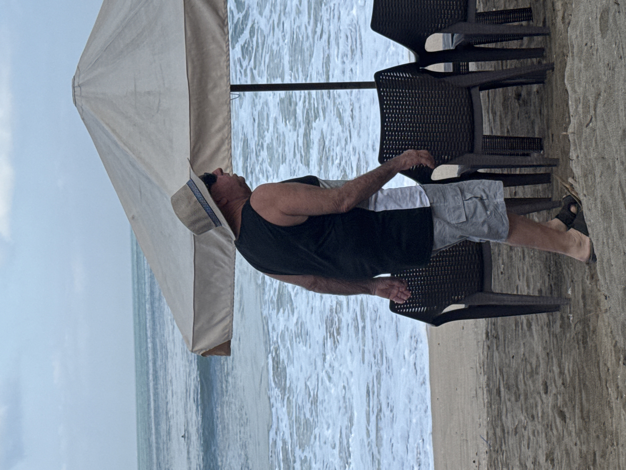 An older Caucasian man wearing a straw hat, sunglasses, a black tank top over a white shirt, and light grey cargo shorts is walking on a sandy beach in Kuta, Indonesia. He is wearing dark sandals. Behind him, there are several dark plastic chairs and a large beige umbrella. The ocean with breaking waves is visible in the background, and the sky is overcast.