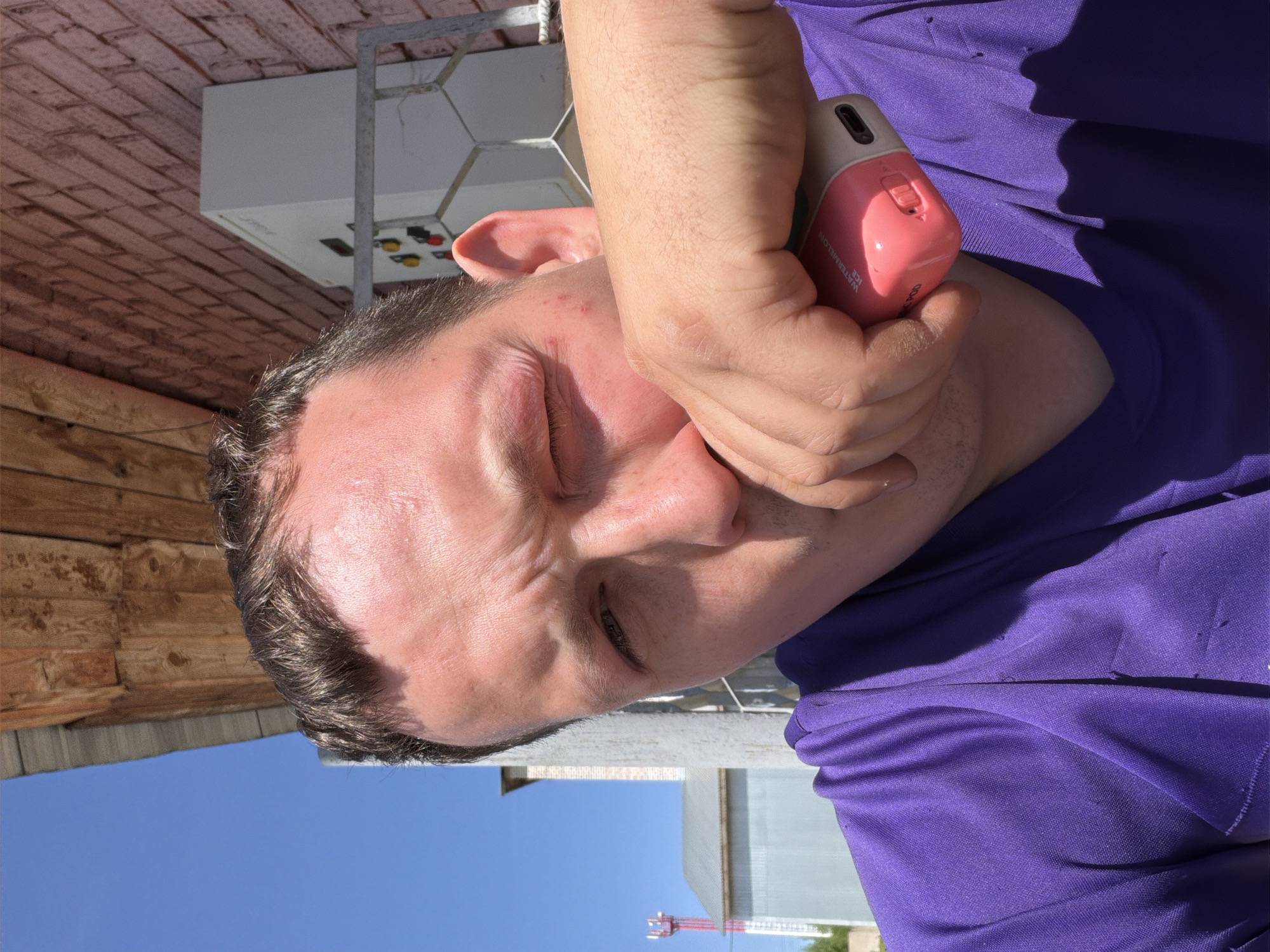 A man in a purple shirt holds a pink vaping device to his face. The location is Chaikovsky, Russia. In the background, there is a building and a communication tower.