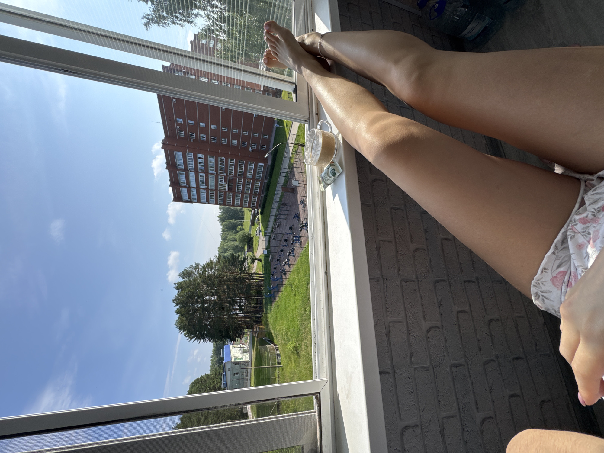 A person's legs are visible, stretched out on a windowsill. In front of the legs, there is a glass mug of coffee and a candy bar. Beyond the windowsill, there is an outdoor sports ground with exercise equipment and trees. In the background, there is a multi-story apartment building. The sky is blue with some clouds. The scene is an outdoor balcony in the city of Chaikovsky, Russia, on a sunny day.
