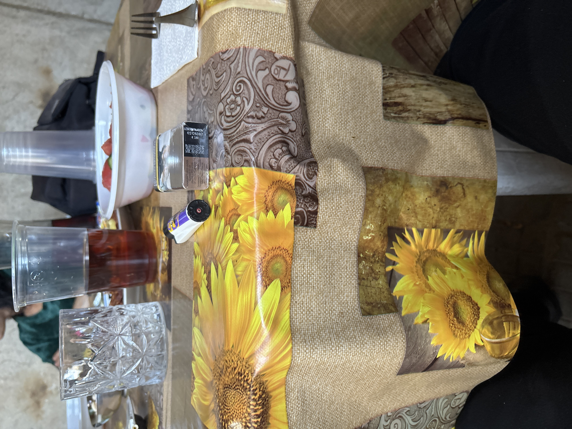 A table covered with a multi-patterned tablecloth is the central focus of the image. The tablecloth features sections with sunflower designs, a woven burlap texture, a wood grain pattern, and an embossed brown floral design. On the table, a white bowl containing what appear to be cut strawberries is visible, along with several clear plastic cups, one filled with a reddish-brown liquid. A crystal glass and a metal fork are also present. A small black box, seemingly a product container, and a small roll of film are also on the table. The background shows glimpses of a person's clothing, suggesting someone is seated at the table. The location is Rakovski, Bulgaria, although the image does not inherently confirm or deny this.