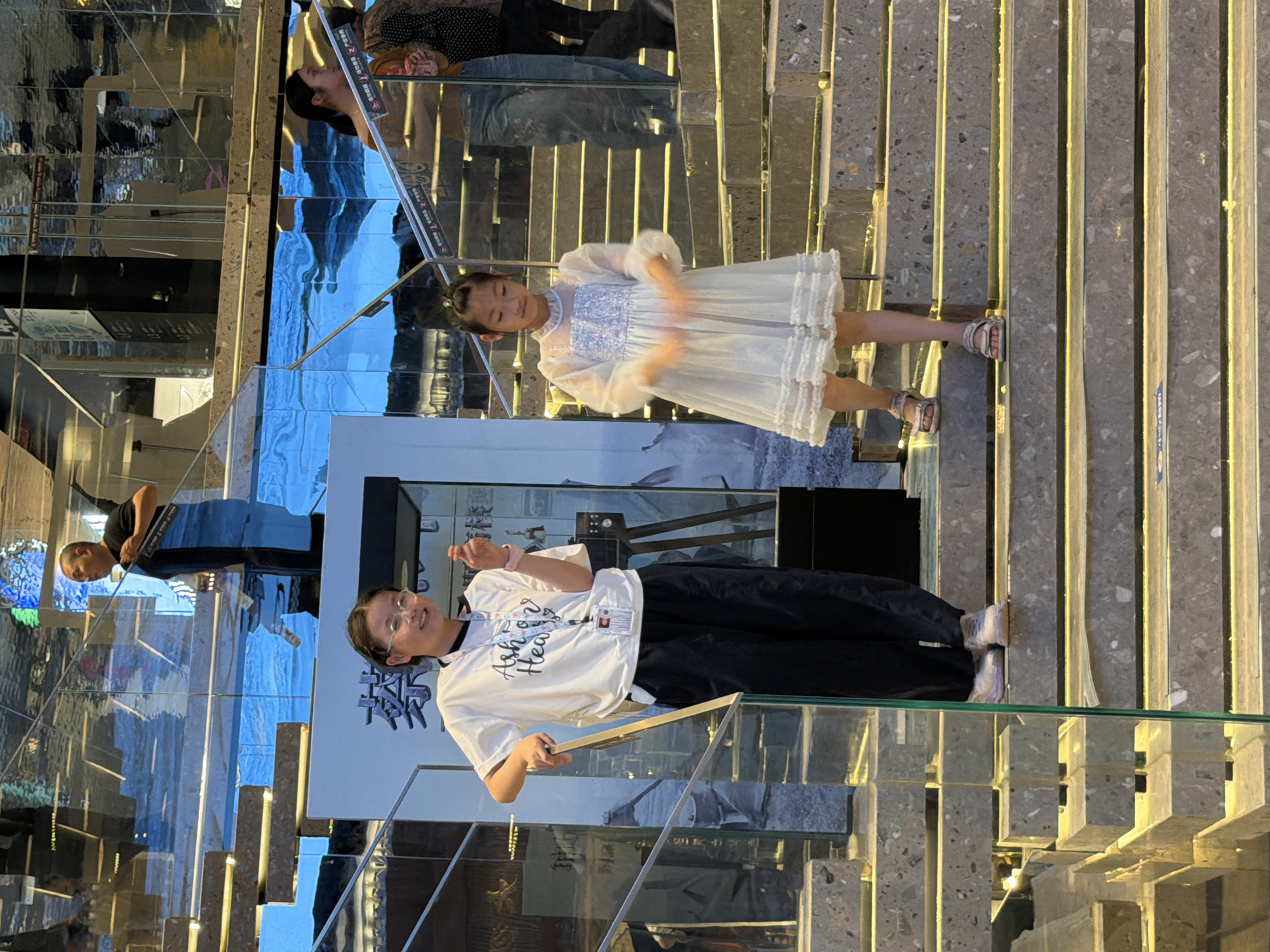 In Yuzhong District, China, two young girls stand on a modern, multi-level staircase. The girl on the left, wearing glasses, a white t-shirt with "Ash Heart" printed on it, and black pants, leans against a glass railing. She is smiling and holding up her right hand, appearing to gesture or interact with something. The girl on the right, in a white, frilly dress, stands with her arms outstretched, her hands blurred as if in motion. Behind them, the staircase features grey stone steps with illuminated yellow strips. The background is a reflective surface, possibly glass or polished metal, which mirrors the environment and shows the blurry figures of other people, including a man in a black shirt standing higher up on the stairs. The overall scene suggests a public indoor space, possibly a museum or exhibition hall, characterized by contemporary architecture.