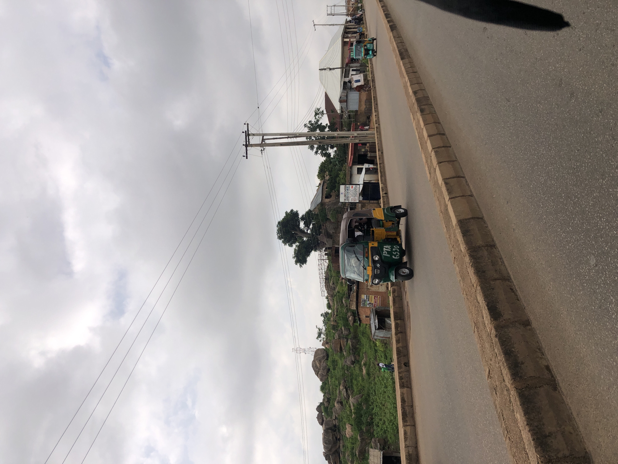 The image shows a street scene in Jos, Nigeria, under a cloudy sky. A green and yellow auto-rickshaw, with the license plate "PTA 6530," is driving on the right side of the road. In the background, there is a rocky, green hillside dotted with trees and small buildings. Power lines stretch across the sky. Another auto-rickshaw, turquoise in color, is visible further down the road. The street is paved with asphalt and bordered by a concrete curb. A person is walking on the sidewalk next to the hillside.