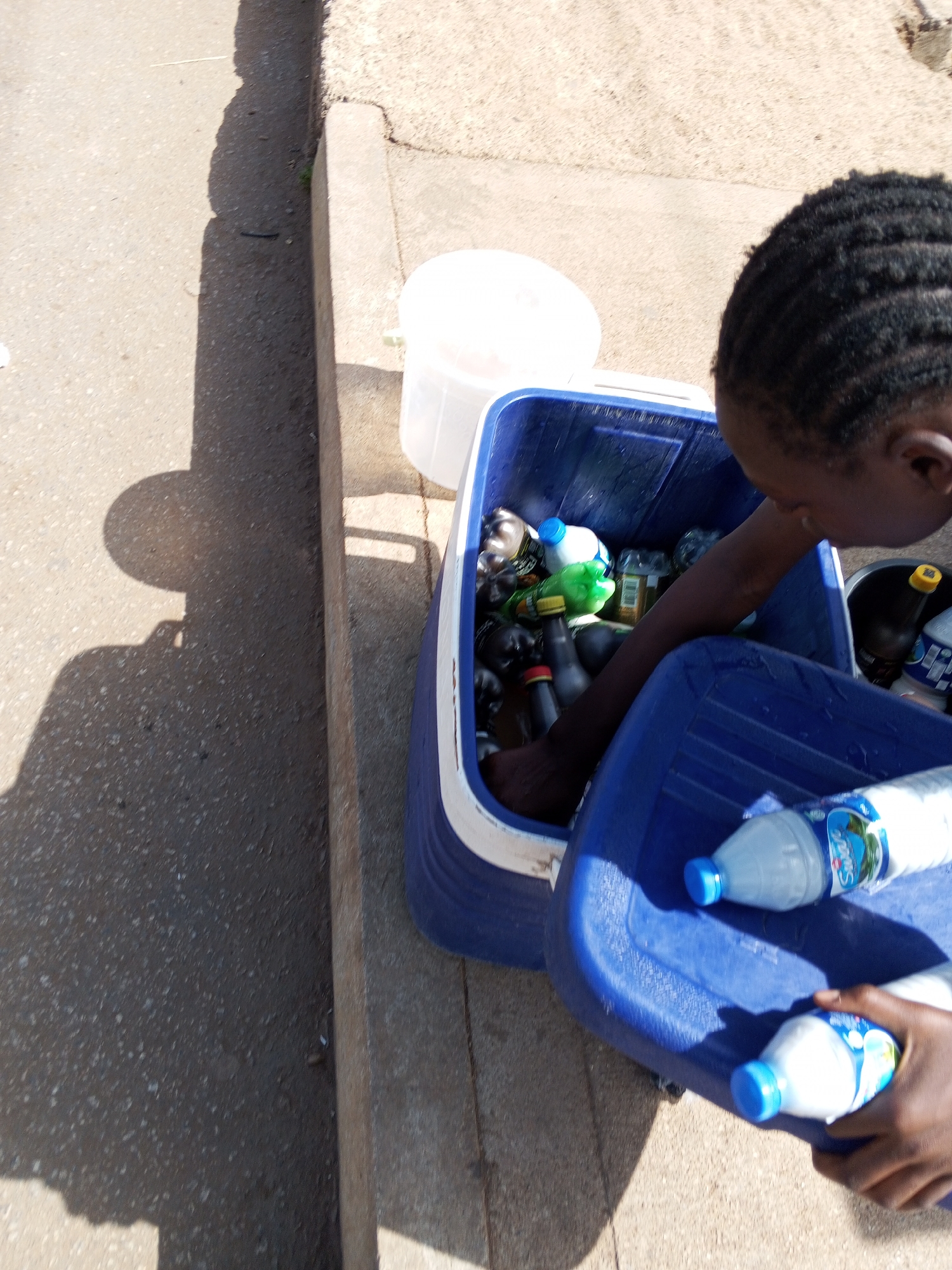A person is sorting through a blue and white cooler filled with plastic bottles. The cooler is placed on a concrete surface. The person is reaching into the cooler with one hand, while the other hand holds a blue lid with two more plastic bottles on it. A white bucket is sitting behind the cooler. The location is Bukuru, Nigeria, based on bottle brands.