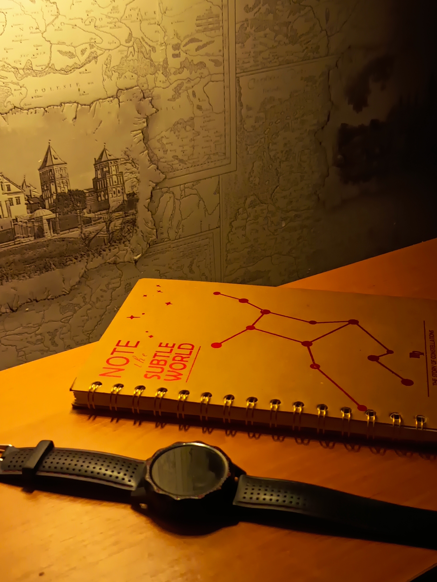 A close-up, low-angle shot shows a black smartwatch with a perforated band lying on a wooden surface in the foreground. To the right, a yellow spiral-bound notebook is open, displaying a red constellation diagram and the text "NOTE the SUBTLE WORLD" and "THE STORY OF CONSTELLATIONS." Behind the notebook and smartwatch, a textured wall serves as a backdrop. The wall is adorned with a vintage-style map on the left, featuring geographical details and buildings resembling old architecture. The lighting is warm and atmospheric, casting shadows and highlighting the objects. There are no people present in the image. The scene does not contain any information that allows for the identification of the city of Izmail, Ukraine.