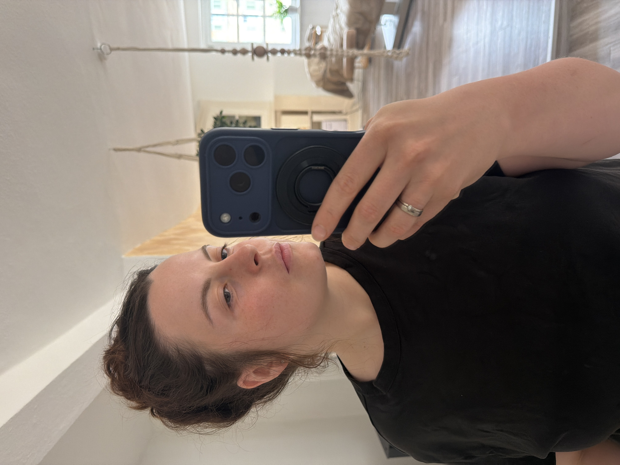 A mirror selfie captures a person holding a dark blue smartphone, revealing both their reflection and the phone's back. The central subject is a person with fair skin and dark brown hair, possibly pulled back, wearing a plain black t-shirt. Their face is visible, looking towards the right side of the frame with a neutral expression.
The person's left hand, adorned with a silver ring on the ring finger, securely holds the smartphone. The phone case is dark blue, featuring a prominent triple-lens camera system on the back, along with a small flash. A black circular pop socket or ring holder is attached to the center of the phone's back, providing a grip.
The background, a reflection within the mirror, shows an indoor setting brightly lit by natural daylight streaming through a large window. Light-colored walls are visible, along with hints of wooden flooring. A decorative hanging element, possibly macrame or a beaded string, is reflected in the upper part of the background. The overall impression is a casual, everyday moment captured indoors during the daytime in what appears to be a home in Meisenheim, Germany. No visible text is clearly legible in the image.