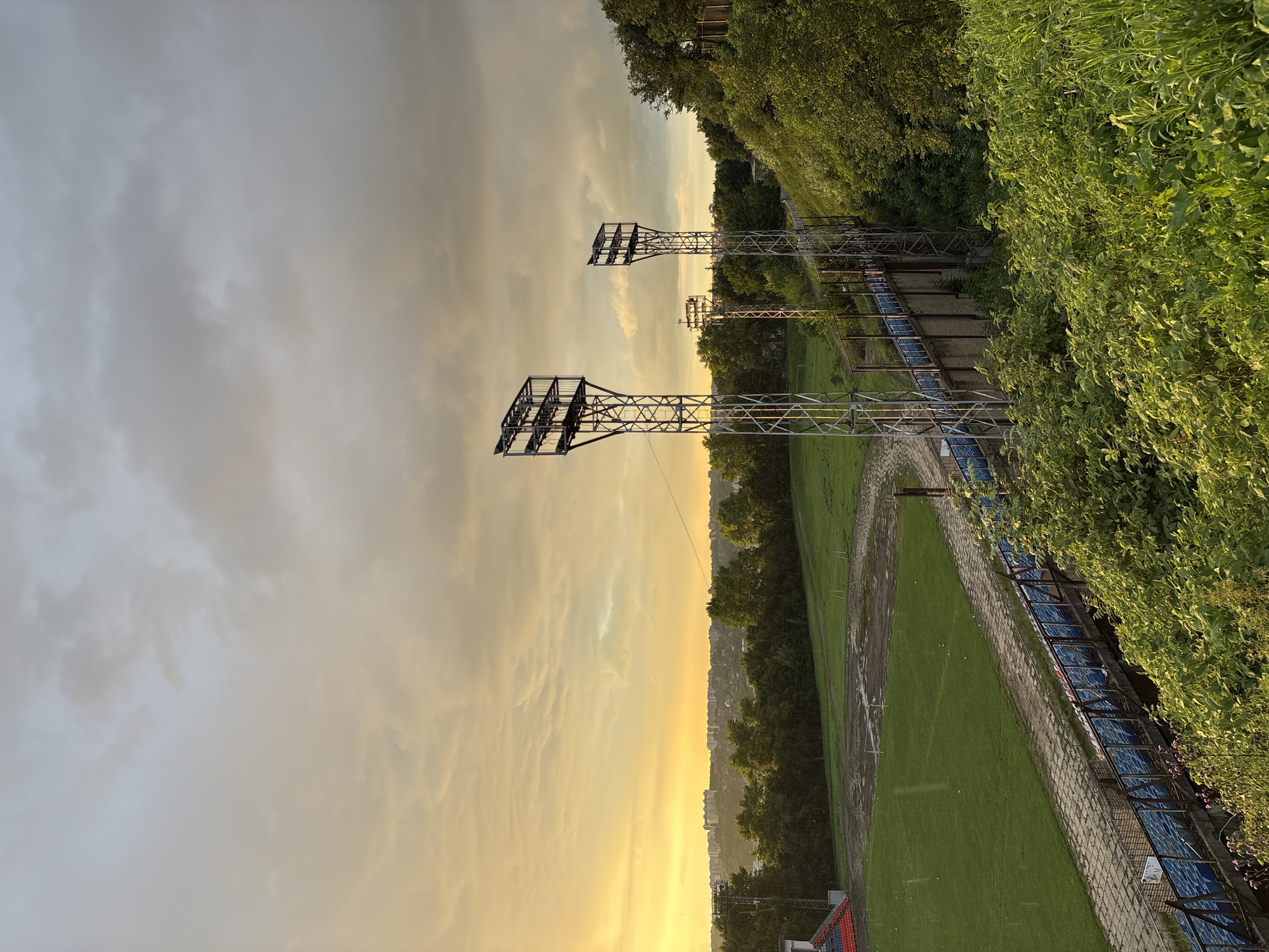 The image depicts an outdoor view of a stadium and its surrounding environment during a cloudy sunset. In the foreground, dense green foliage covers a slope. Behind the foliage, rows of blue stadium seats are visible, separated by metal railings and a concrete walkway. A stadium field, partially wet and marked with white lines, is located to the left of the seats, with a running track encircling it.
Dominating the mid-ground are two tall, metallic floodlight towers with multiple lamps, positioned on a concrete structure. Further in the background, a line of lush green trees separates the stadium from a cityscape with numerous buildings, silhouetted against the glowing orange and yellow sky. The sky itself is filled with dramatic, overcast clouds, with the sun's warm light breaking through. The overall scene suggests an abandoned or less-maintained sports facility under a moody, twilight sky. There are no people visible in the image. The location is identified as Sof'ino, Russia.