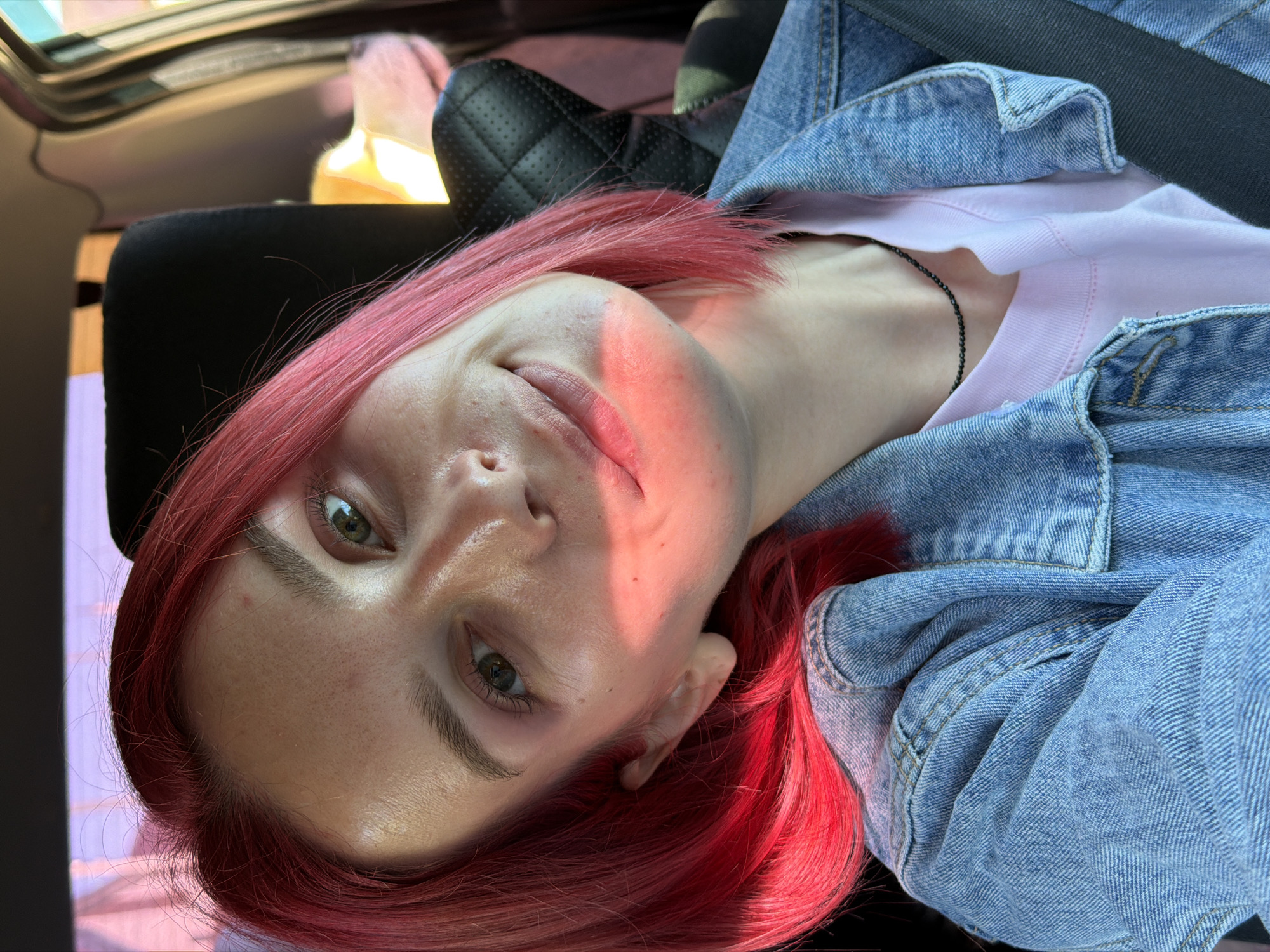 A young woman with bright pink hair is looking at the camera while sitting in a car. She is wearing a denim jacket over a light pink t-shirt and a thin black necklace. The sunlight is hitting her face, creating a warm glow. The background shows a car interior with a black headrest. The overall impression is a casual and natural selfie taken during the day, likely in Chaikovsky, Russia, given the context of the request.