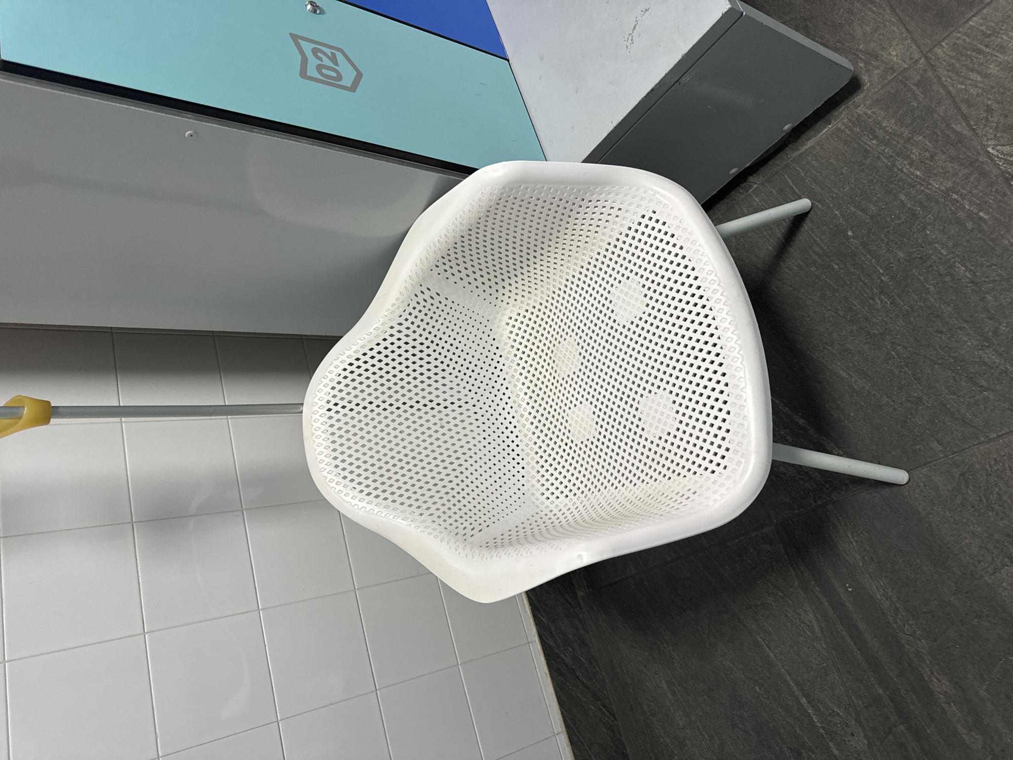 This media file shows an overhead view of a white, perforated plastic chair with a slightly curved seat and back. The chair is positioned on a dark gray, textured floor that appears to be made of large tiles or planks. To the left of the chair, a section of a wall covered in white square tiles is visible, and a metallic pole with a yellow handle extends into the frame from the left. Behind the chair, there is a light blue panel with a grey outline of the number "02" inside a box. A dark gray vertical structure is also present behind the chair, to the right of the blue panel.
The scene appears to be indoors, possibly in a public waiting area, locker room, or similar facility. There is no indication of the time of day or weather conditions. The overall impression is clean and modern. The chair has a contemporary design, and the perforated pattern suggests it might be suitable for outdoor or wet environments, or simply for ventilation. The presence of the tiled wall and the locker-like panel implies a functional space. There are no people visible in the image. The only visible text is the numeral "02" on the blue panel. The context of Murcia, Spain, does not provide specific visual cues within the image itself.