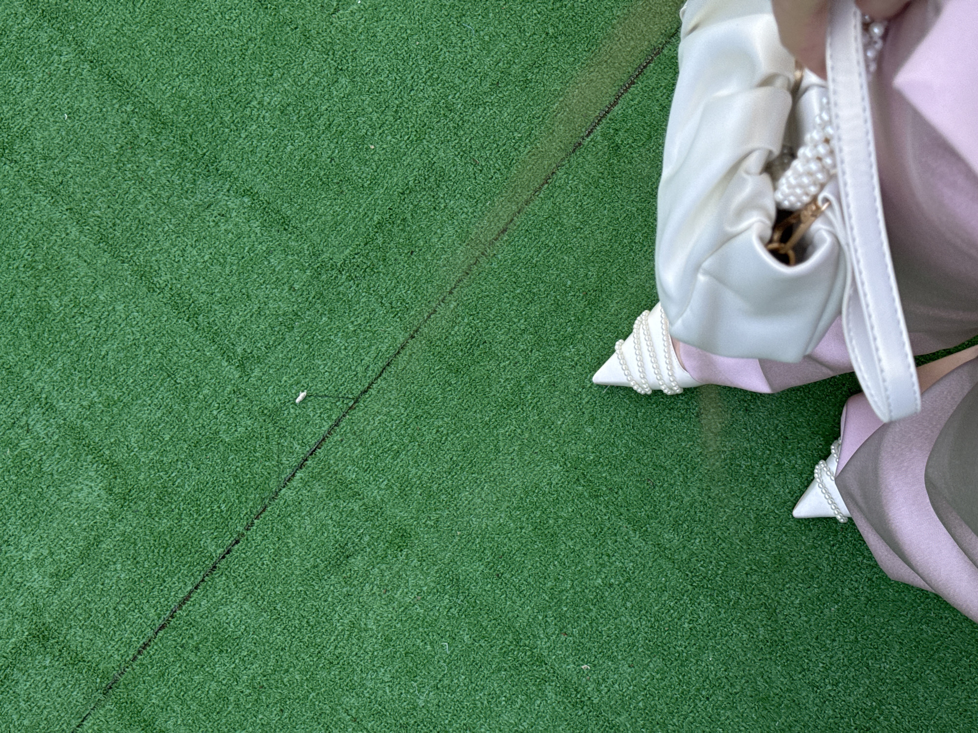 The image shows a close-up, high-angle view of a person's legs and feet dressed in a light pink outfit and white pointed-toe heels adorned with pearls. The person is holding a white, ruched clutch bag with a pearl embellishment. The background is artificial green turf with a faint dark line running diagonally across it. The scene does not provide any discernible details that would indicate a location in Moscow, Russia.