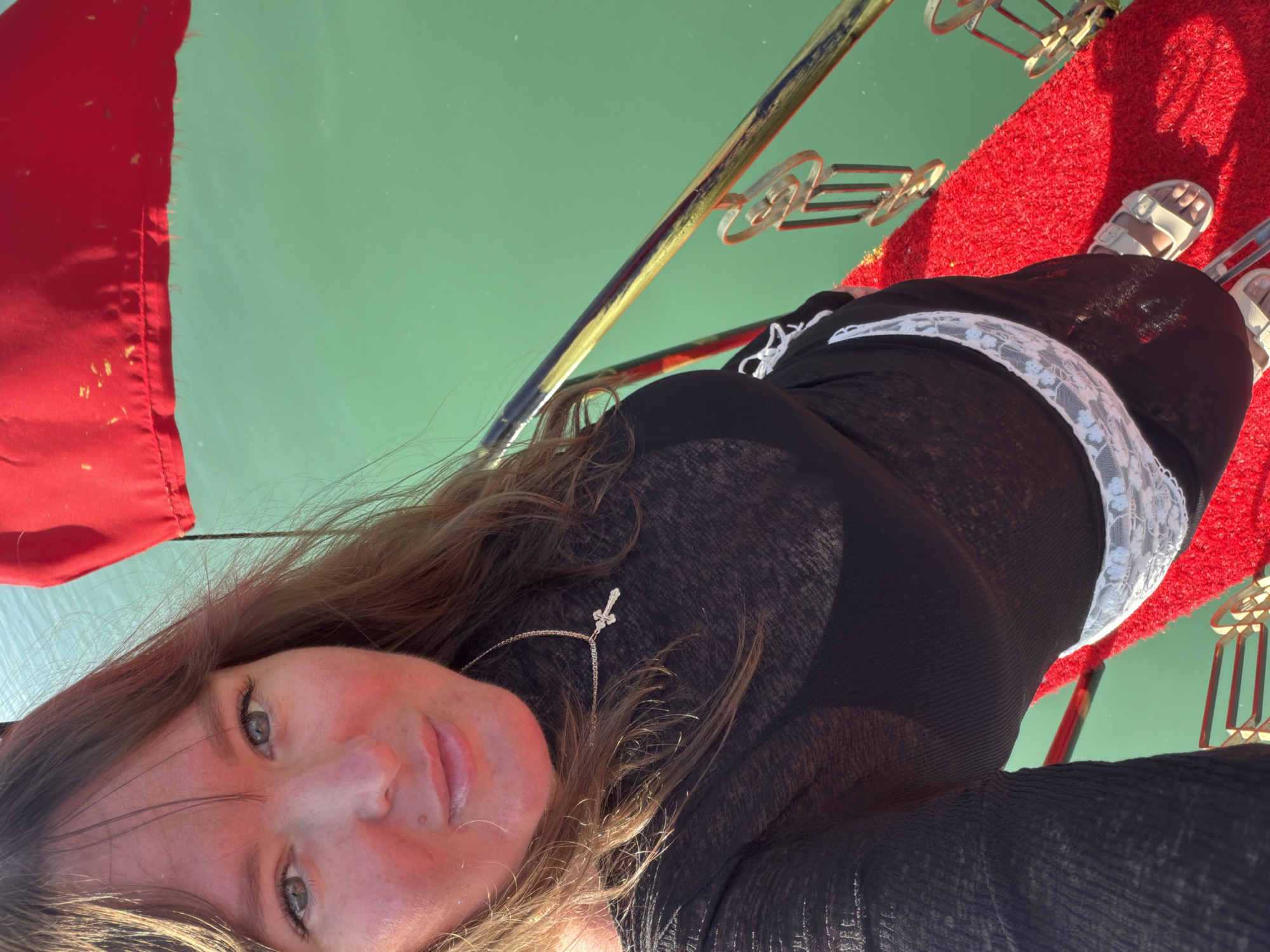 The photograph appears to have been taken from a first-person perspective, with the subject seemingly lying down on a red surface, possibly a carpet, aboard a vessel. The vessel seems to be navigating turquoise water, which may suggest it is on a river or a coastal area. A red flag is visible on the left side of the frame, potentially indicating the vessel is affiliated with a specific entity or region. The subject is wearing a black dress and white sandals. The surrounding area includes decorative metal elements, adding to the vessel's ambiance. Based on the presence of a Turkish flag and the color of the water, the photograph may have been taken during a trip to Alanya, Turkey.