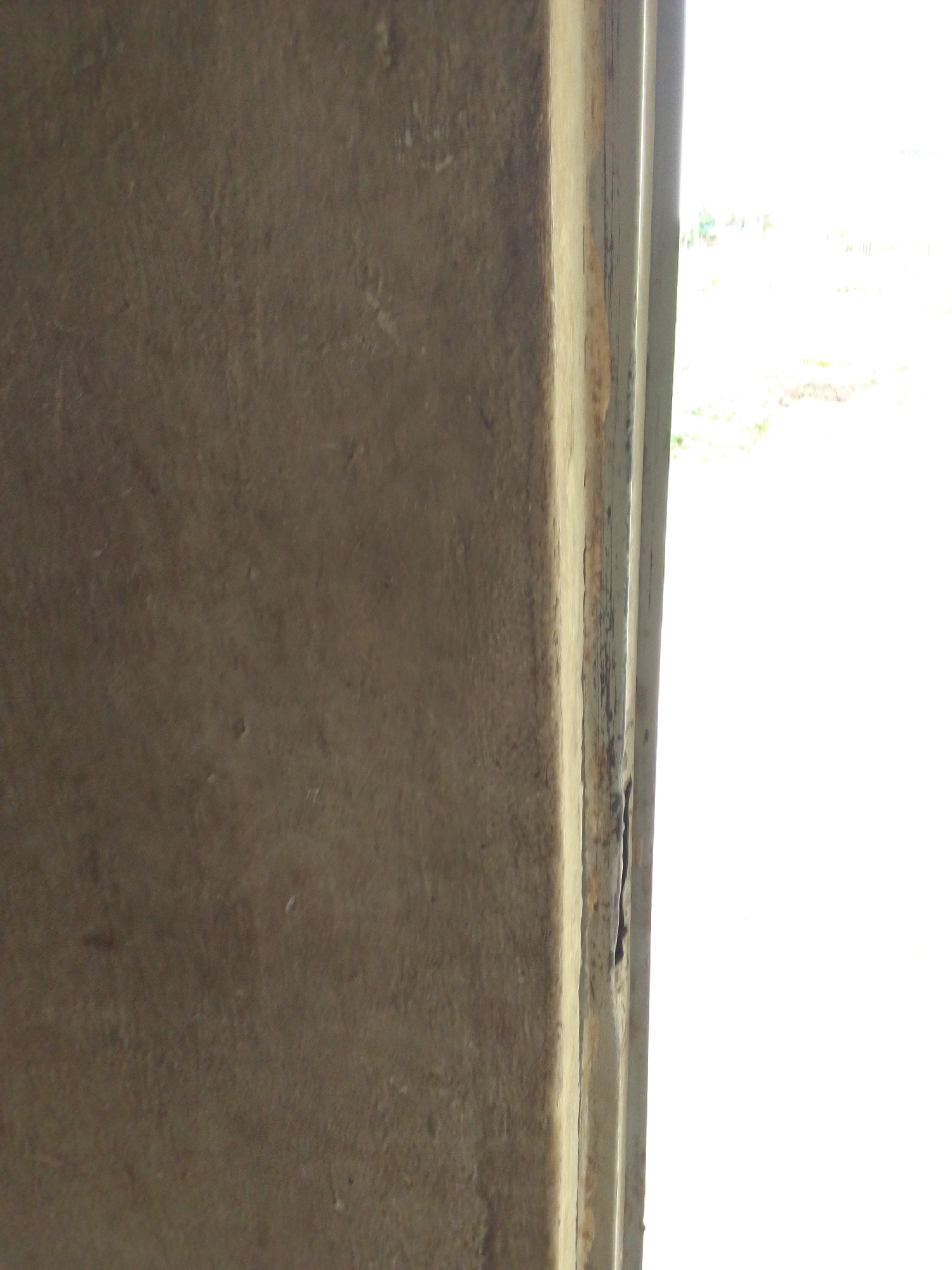 The image shows a close-up, slightly angled view of a weathered, textured concrete wall. A thin, vertical strip of what appears to be a wooden or metal frame, painted a light beige with darker, weathered sections, runs along the right side of the concrete wall. This frame creates a slight shadow and a visual separation between the wall and the bright, overexposed background on the far right. The background is indistinct, suggesting an outdoor, brightly lit environment, possibly with some faint greenery or natural elements visible. The overall impression is one of rough texture and aged materials.