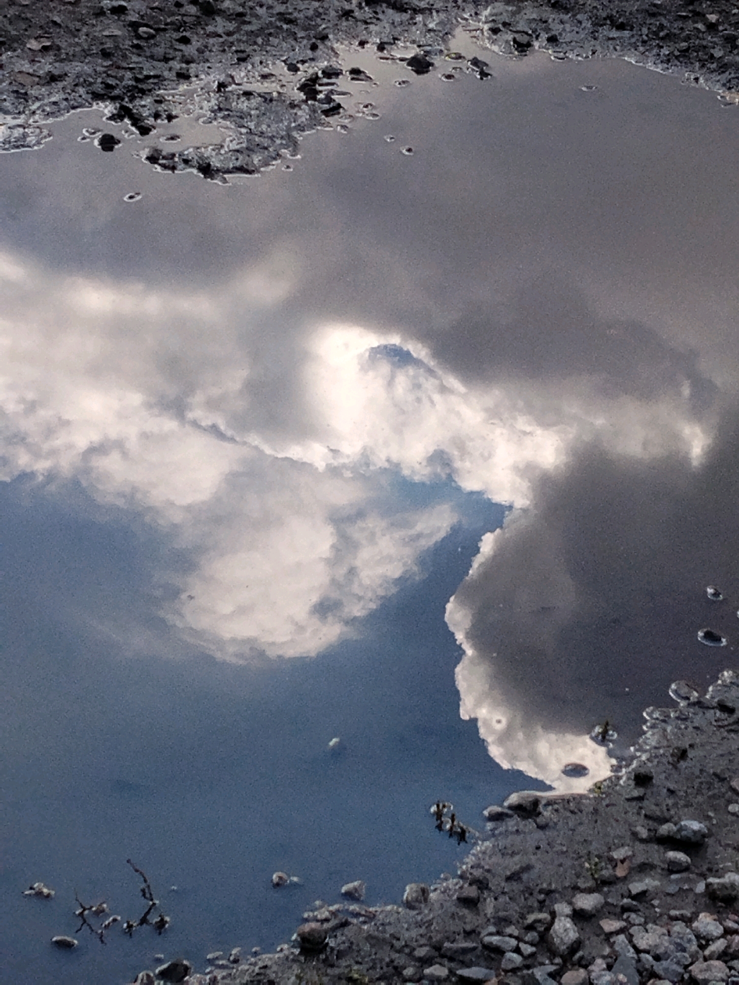 The image displays a reflection of clouds in a puddle of water on a rocky surface. The sky is filled with dramatic, heavy clouds, with some areas of brighter white suggesting sunlight breaking through. The water in the puddle is dark and reflective, mirroring the cloudy sky with striking detail. The edges of the puddle are lined with rough, gray stones and small pebbles, indicating an outdoor environment, possibly a street or unpaved area. There are no visible people or animals in the frame. The scene appears to be an abstract composition of natural elements captured in a moment of reflection. The location in Hoholeve, Ukraine, is noted but not visually represented within the image itself; the image only captures the puddle and its reflection.