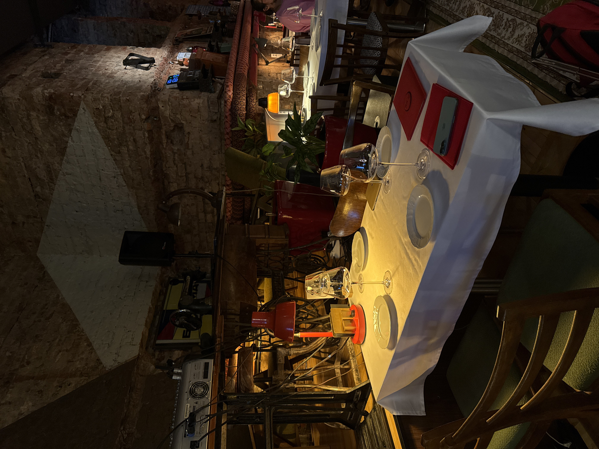 This image shows a restaurant in Moscow, Russia. The restaurant has a rustic aesthetic, with exposed brick walls. There are tables set with white tablecloths, plates, wine glasses, and red placemats. Some tables are adorned with plants in red pots. A smartphone rests on one of the placemats. There's an array of vintage equipment, including a speaker, a car racing game, and assorted electrical equipment. The lighting is dim and warm. Some people are seated at tables in the background.