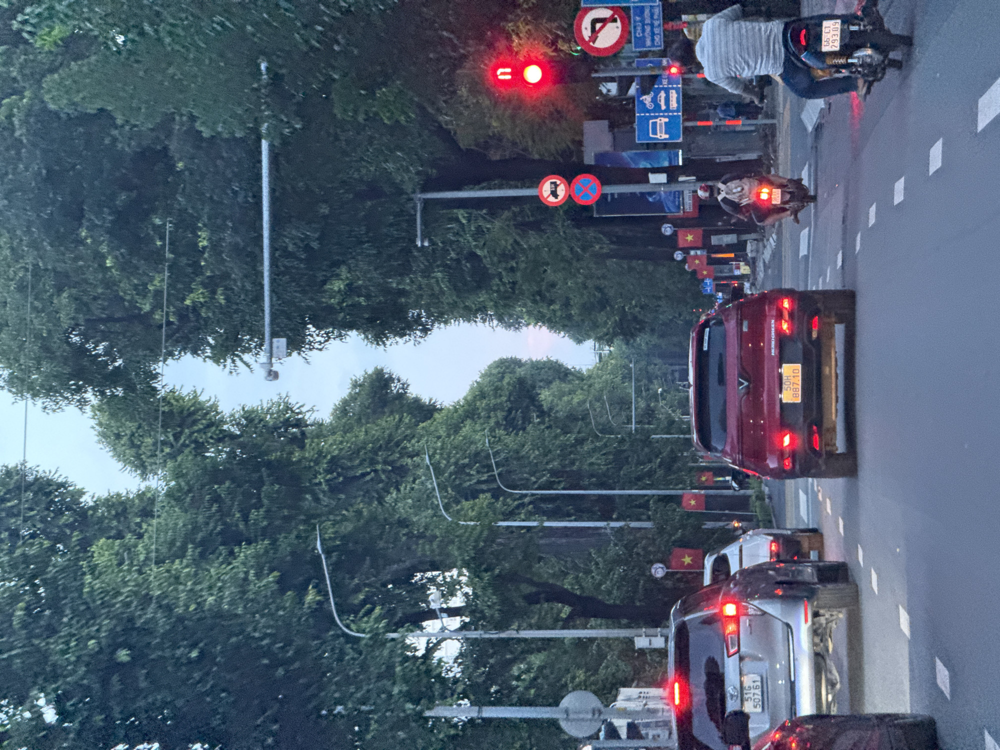 The image shows a street scene in Thủ Đức, Vietnam, likely during the daytime. A line of cars and motorcycles are stopped at a traffic light, indicating traffic congestion. The road is lined with tall, mature trees that form a canopy over the street. Traffic lights are visible, with one showing red. Several traffic signs are present, including a "no stopping" sign and a sign indicating a right turn is permitted. Vietnamese flags are also visible, suggesting a national holiday or event. The atmosphere appears to be calm, with no visible signs of emergency or distress.
