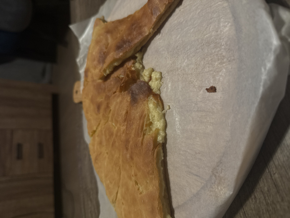 A golden-brown pie, possibly khachapuri, rests on white parchment paper. The crust is uneven and slightly charred in places, revealing a creamy, pale filling along the exposed edge. A slice has been removed from the top. A small crumb rests on the paper to the right. The scene is set on a wood surface, and a cabinet is partially visible in the background.