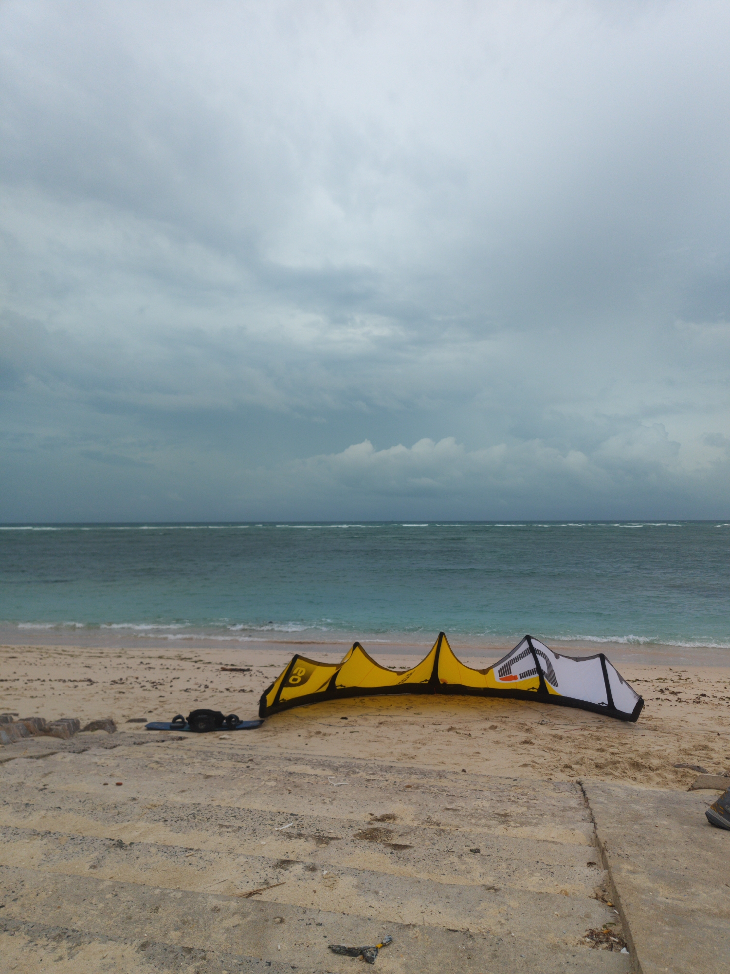 The image captures a tranquil coastal scene in Phú Quý Special Administrative Region, Vietnam, under an overcast sky. The main subject is a large kiteboarding kite, predominantly yellow and white with black accents, lying deflated on a sandy beach. Adjacent to the kite, a black kiteboard with bindings and a control bar are also visible on the sand.
The scene depicts a calm sandy beach with light-colored sand, leading into clear, turquoise-to-dark-blue ocean waters. Gentle waves break near the shore, and further out, lines of larger waves indicate a reef or shallows. The horizon is visible where the sea meets the cloudy sky. The sky is filled with heavy, gray clouds, suggesting an overcast day, possibly before or after rain, or simply a consistently cloudy day, making it difficult to pinpoint the exact time of day beyond daylight.
In the foreground, a concrete structure, possibly a path or steps, separates a higher sandy area from the immediate beach where the kite and board rest. No people are visible, implying a moment of rest, preparation, or quiet observation of the equipment before or after use.
Notable details on the kite include visible text: on the yellow section, the letters "CO" are discernible, likely part of the brand name "CORE." On the white section of the kite, a black graphic logo is present, and beneath it, the letters "XR" are visible, identifying it as a "CORE XR" model kite. The overall atmosphere is one of peaceful anticipation or quiet solitude by the sea.