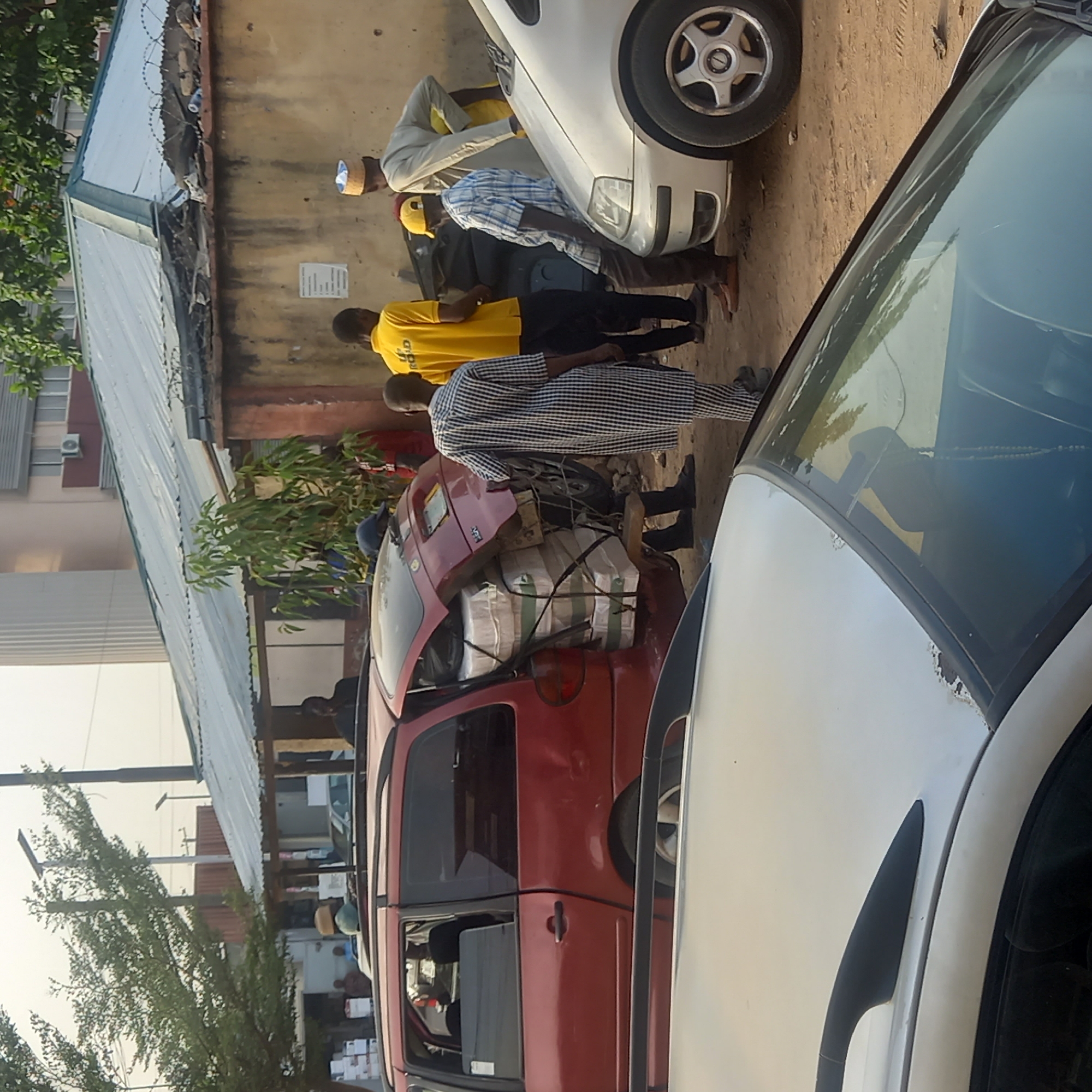 This image captures an outdoor scene in Dadi Riba, Nigeria, during daylight hours under sunny weather. Several men are gathered around two parked cars: a white sedan and a red hatchback.
The central focus is a red hatchback car, which is extensively loaded with numerous white packages or sacks. These items are secured tightly to the car's roof and visible through its open or damaged rear window with green and white ropes. A man wearing a black and white checkered traditional tunic and dark shoes stands near the driver's side of this heavily loaded red car.
Closer to the white sedan, another man in a yellow t-shirt, with the word "ROLD" partially visible on it, stands facing away from the camera. Beside the front of the white car, a man in a light-colored traditional outfit and a woven cap is also present. Other individuals are visible in the background, suggesting a busy street or commercial area.
The ground beneath the vehicles and people is unpaved, appearing as dirt or sand. In the background, there are buildings with corrugated metal roofs, one of which features barbed wire on top of its wall. Green foliage from trees and bushes is seen on the left and central parts of the image, contributing to the outdoor setting. A small, unreadable white sign with black text is affixed to the wall in the background. No strong emotions are discernible from the individuals; they appear to be engaged in or observing a task related to the vehicles and packages.
