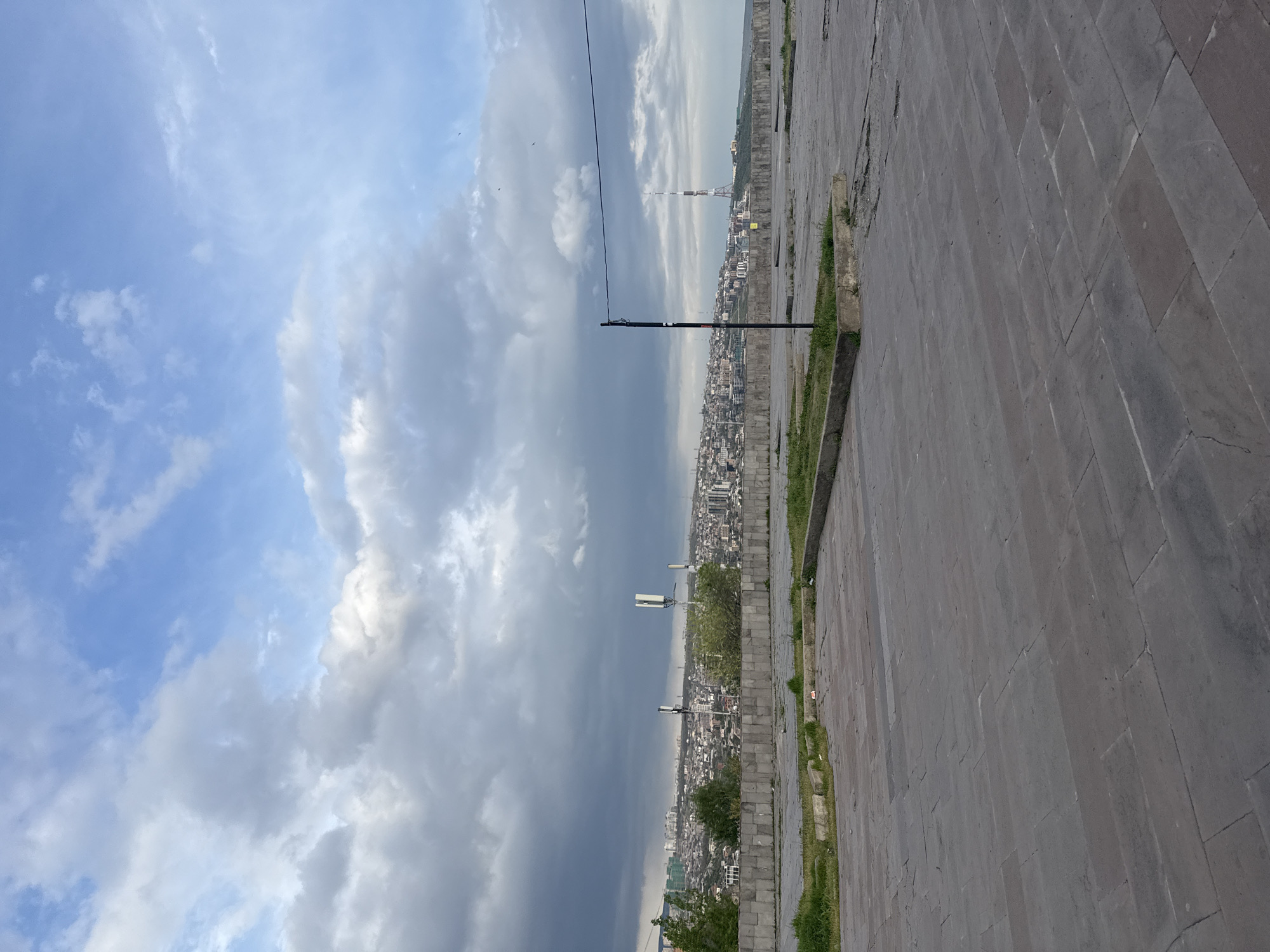A panoramic daytime view overlooks a vast urban landscape, identified as Yerevan, Armenia, from an elevated vantage point. The sky dominates the upper portion of the frame, showcasing a dramatic contrast between bright blue patches dotted with white, wispy clouds and large expanses of heavy, dark grey clouds, indicating dynamic weather conditions.
In the mid-ground, the sprawling city unfolds with a dense arrangement of buildings extending towards the horizon. A tall, distinctive communication tower, consistent with the Yerevan TV Tower, is visible in the distance, rising above the urban sprawl. Along the edge of the elevated foreground, several utility poles or streetlights are positioned, fitted with multiple white rectangular antennas, offering a clear view of the city below. A thin black wire stretches horizontally across the sky, originating from one of these poles.
The foreground consists of a wide paved area composed of large, rectangular, dark grey stone blocks, exhibiting some signs of cracks and weathering. A narrow strip of vibrant green grass grows along the edges of this paved surface, adding a touch of nature to the constructed environment. The lighting suggests it is daytime, likely late morning or afternoon. No individuals are visible, and there is no apparent activity taking place. There is no visible text in the image.