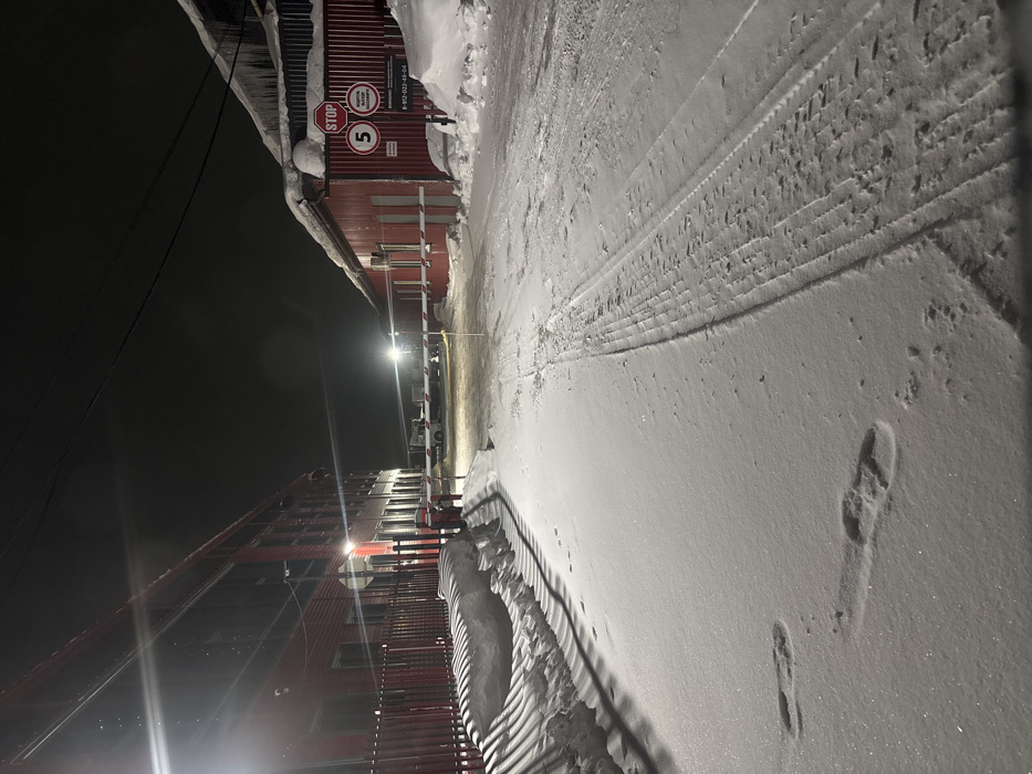 The image depicts an illuminated, snow-covered industrial or commercial area at night in Kamyennoye, Russia. The ground is uniformly covered in fresh snow, exhibiting distinct tire tracks running parallel on the right side of a central path and several individual footprints on the left side. To the left, a two-story building with red corrugated siding and multiple illuminated windows stands behind a red metal fence, paralleling the path. A large snowdrift with ridged shadows lies between the fence and the path. To the right, a single-story building, also with red corrugated siding, features a heavily snow-laden roof with prominent icicles. Attached to a post near this building are a red octagonal "STOP" sign, a white circular "5" (speed limit) sign, and a rectangular sign with Russian text, including "ВНИМАНИЕ ОПАСНО!" (Attention: Dangerous!) and a phone number "8-912-022-48-04". A white and red striped barrier arm extends across the path, indicating a controlled access point. Beyond the barrier, a light-colored vehicle is dimly visible in the distance, illuminated by a further streetlight. Overhead power lines traverse the dark night sky. No individuals are visible.