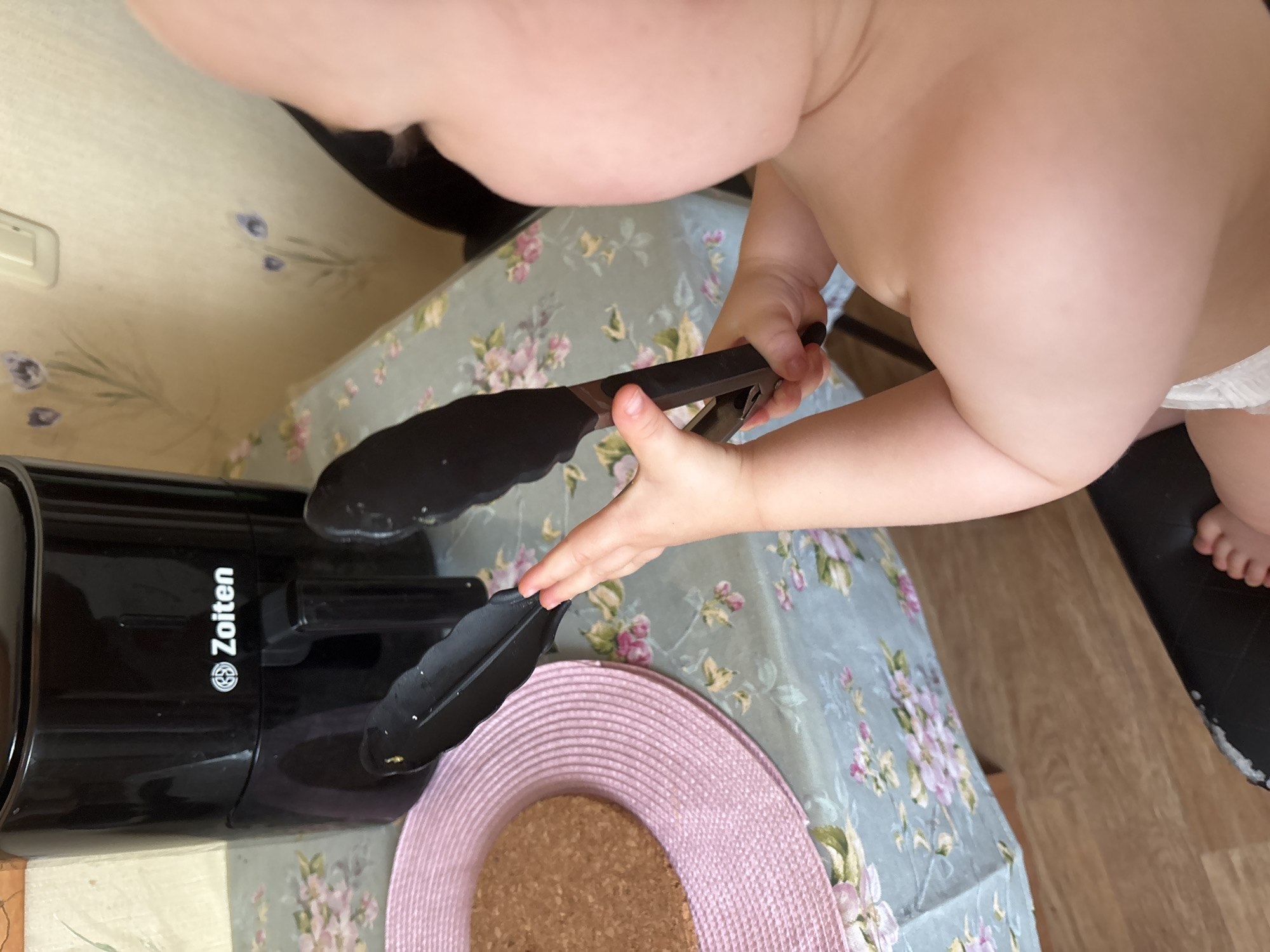 An infant is held up by the arm, as if by an unseen adult, with a black kitchen tong in its hands. The child is wearing a diaper and has fair skin. The tong is black with a metal hinge. The background shows a black air fryer with "Zoiten" logo on it and a pink placemat with a brown circular object on it. The table is covered with a tablecloth with floral patterns. The room appears to be a kitchen. There is no visible indication of the city of Naberezhnye Chelny, Russia.