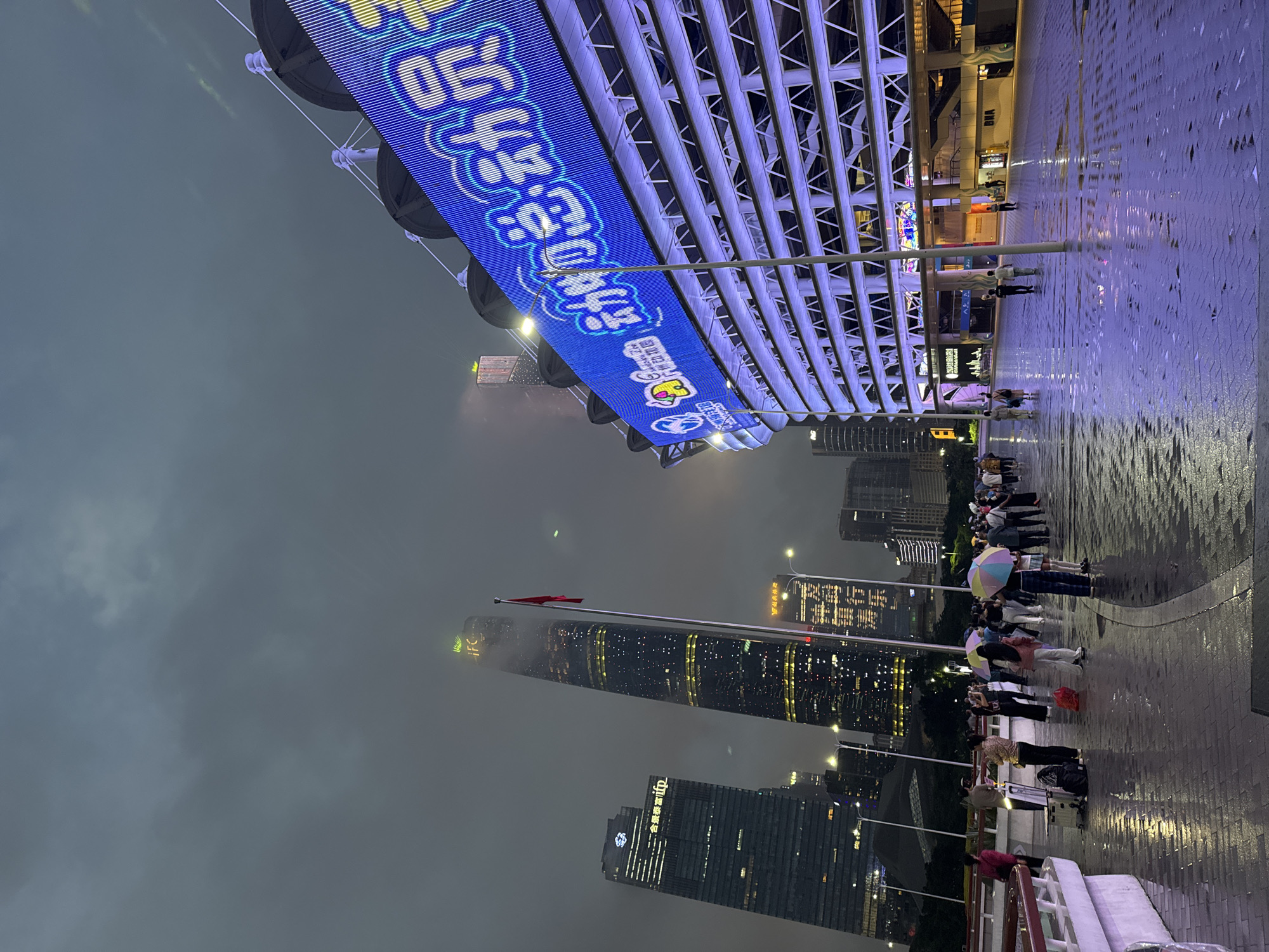 The image shows an elevated pedestrian walkway in Tianhe District, China, at night. It is raining, and the ground is wet. People are walking along the walkway, some holding umbrellas. In the background are tall buildings, including the Guangzhou International Finance Center (IFC), partially obscured by fog or clouds. A Chinese flag flies on a pole near the IFC. A large electronic billboard with Chinese characters is mounted on the structure of the walkway.