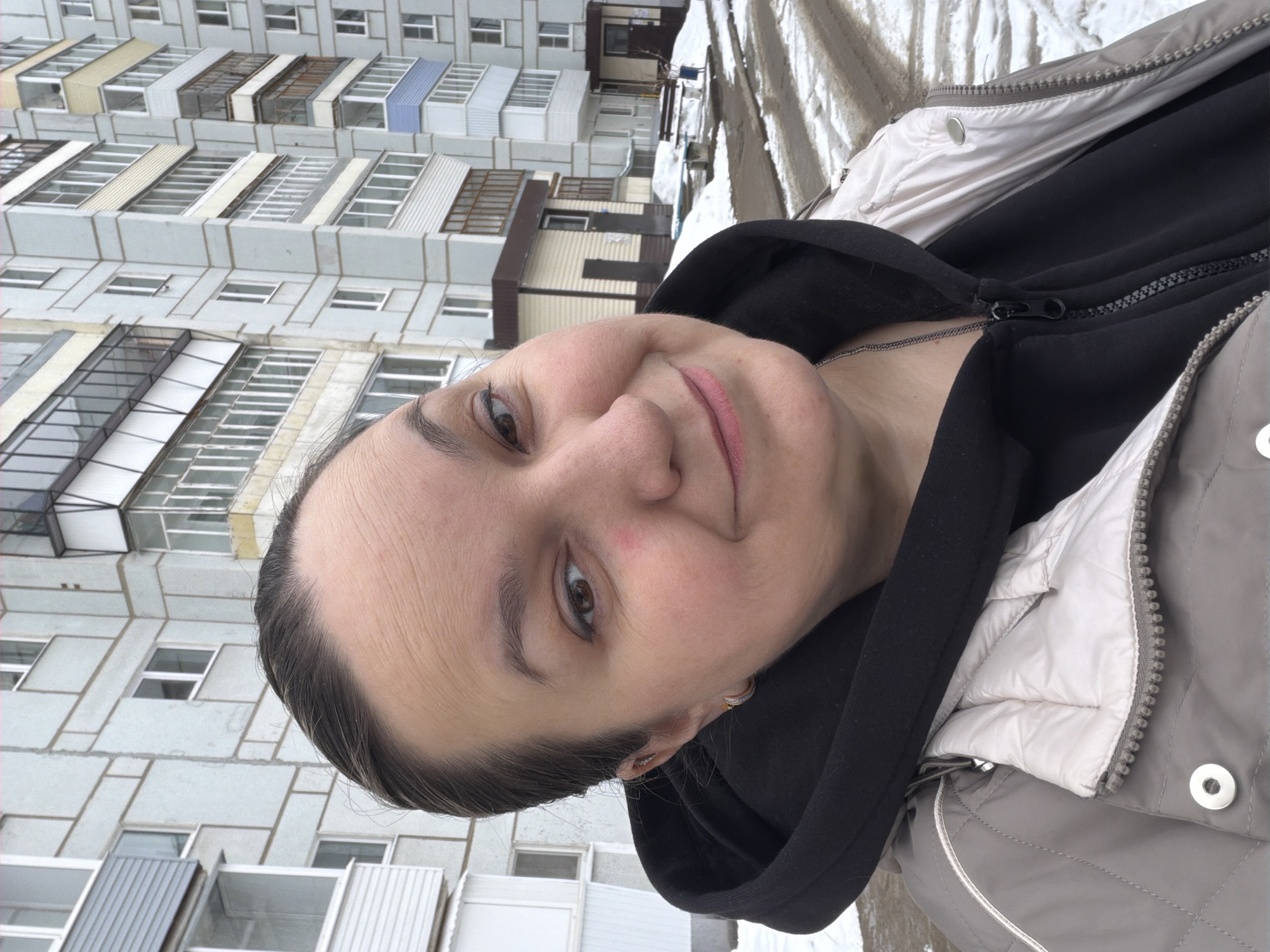 A woman is taking a selfie outdoors, with a multi-story apartment building in the background. The building has multiple balconies, some with different colored panels. The woman is wearing a black hooded sweatshirt and a gray jacket. The environment appears to be cold, with patches of snow visible. Based on visual markers (style of building, general environment) the image is likely taken in Chaikovsky, Russia.