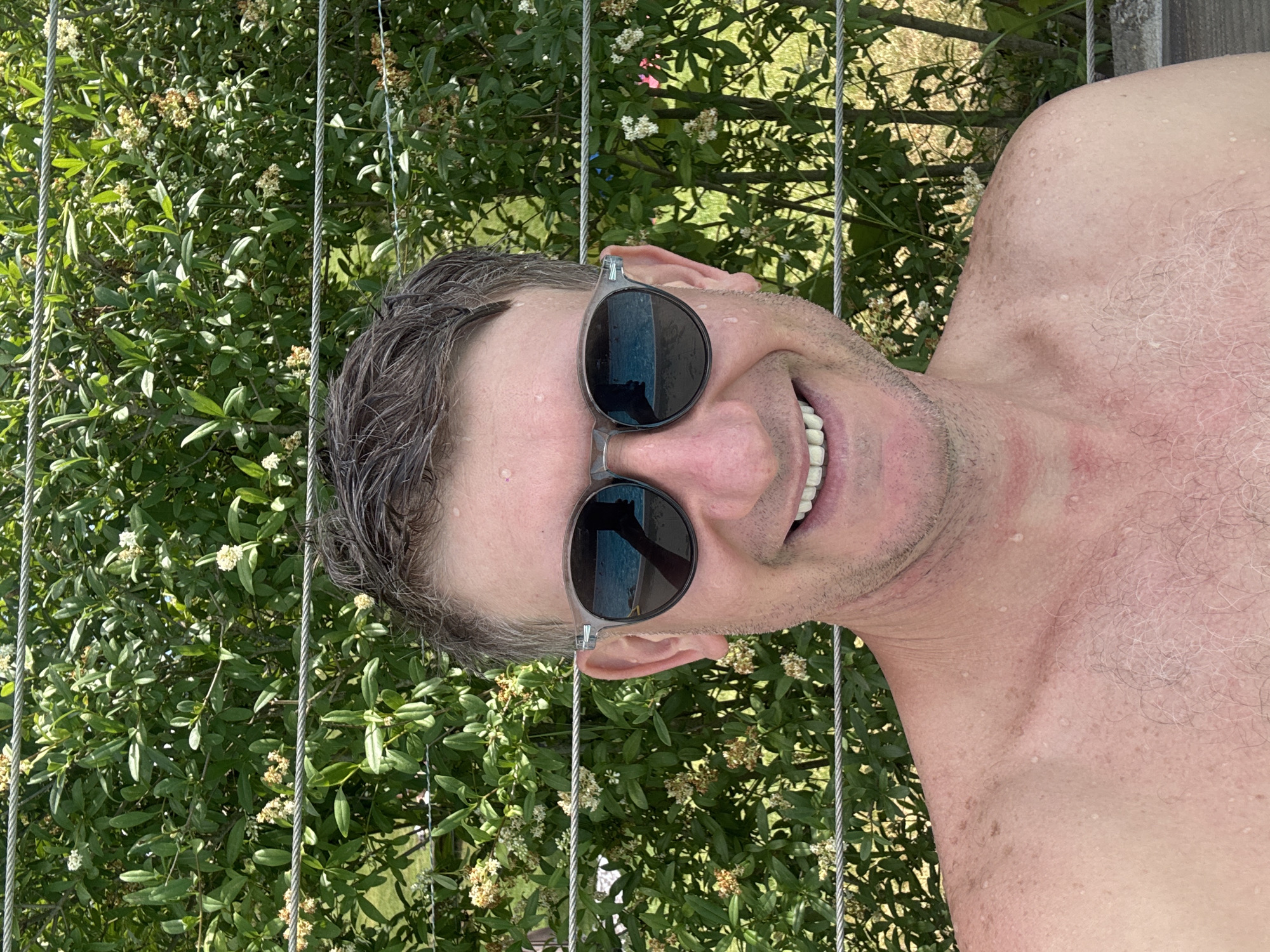 A close-up, low-angle selfie shows a person from the chest up, with their head tilted back and facing the camera. They are wearing dark, round sunglasses and have wet, dark hair. The person is smiling broadly, revealing their teeth. Their skin is pink and appears to have water droplets on it, suggesting they may have been swimming or are in a humid environment.
The background is dominated by lush green foliage, with some small white flowers visible. Thin, metallic wires run vertically through the frame, partially obscuring the greenery. The lighting suggests it is daytime, possibly sunny. The overall mood is relaxed and happy.
The image was taken in Saint-Imier, Switzerland. There is no visible text in the image.