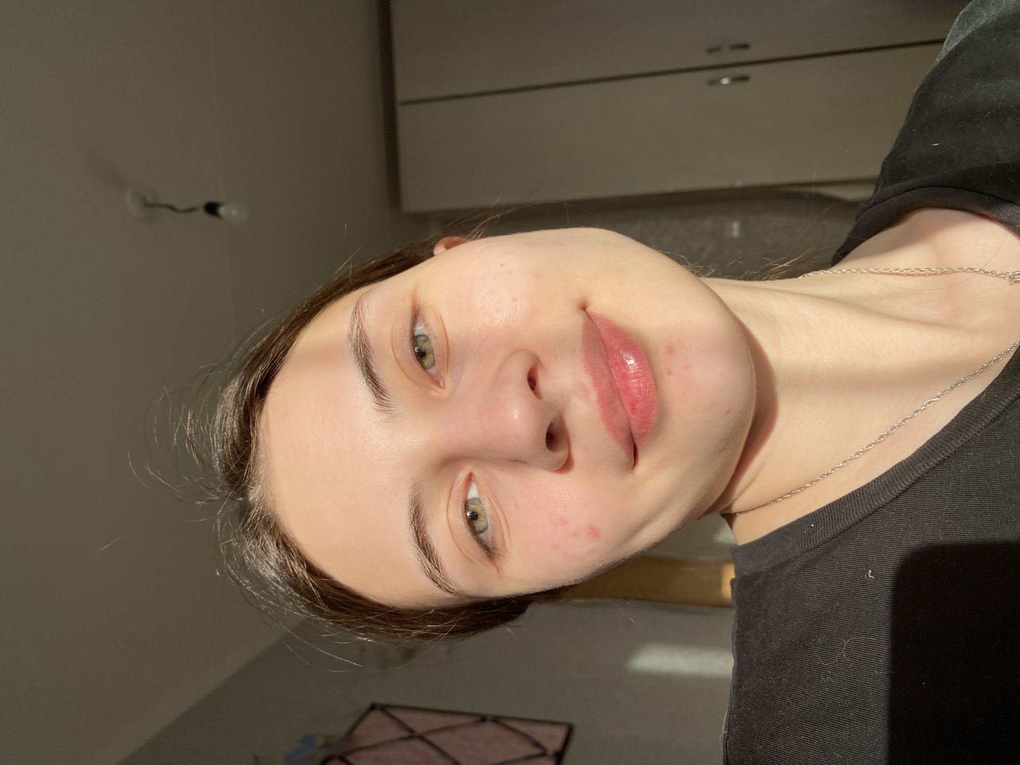 A close-up portrait of a fair-skinned young woman with brown hair and green eyes. She is wearing a black top and a silver necklace. The background includes a white wall, a light fixture, and a cabinet. The setting is not identifiable as Votkinsk, Russia, based solely on the visual information in the image.