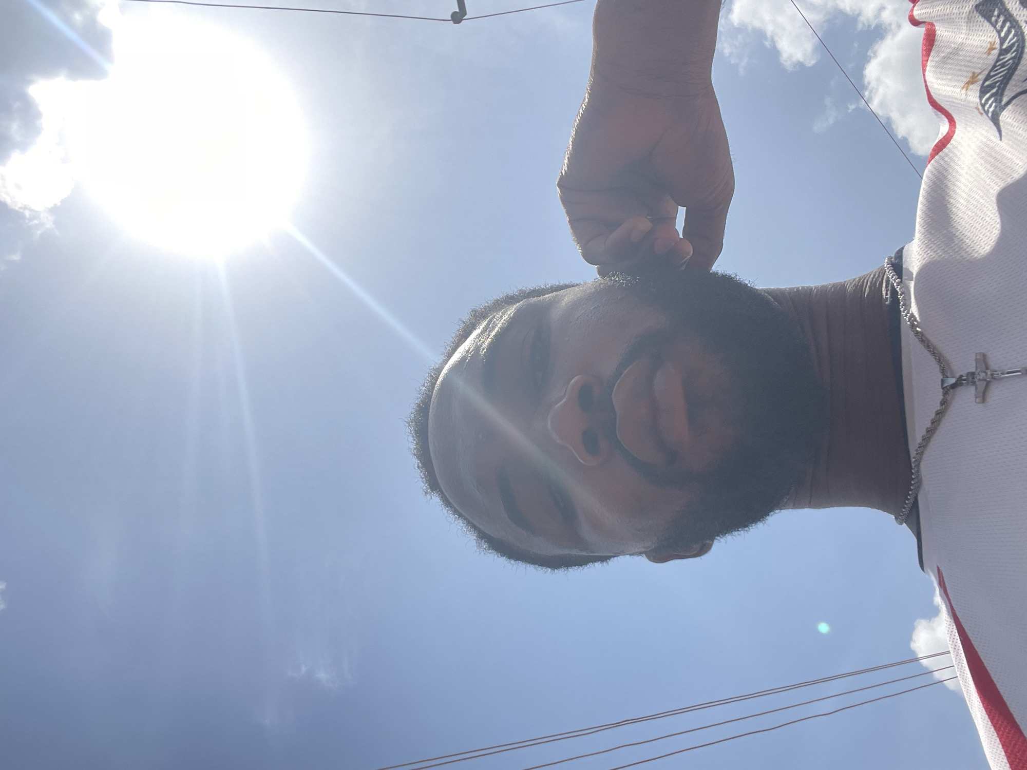 A low-angle, outdoor shot shows a Black man's face close to the camera, his fingers touching his hair. He has a beard. The bright sun is in the upper left of the frame, creating a glare. Above and surrounding him is a clear blue sky with scattered clouds. In the lower part of the image, electrical wires stretch across the sky. He is wearing a white athletic jersey with red and black trim and a silver necklace with a cross pendant. The location is possibly Lokoja, Nigeria, though there is no specific identifying marker.
