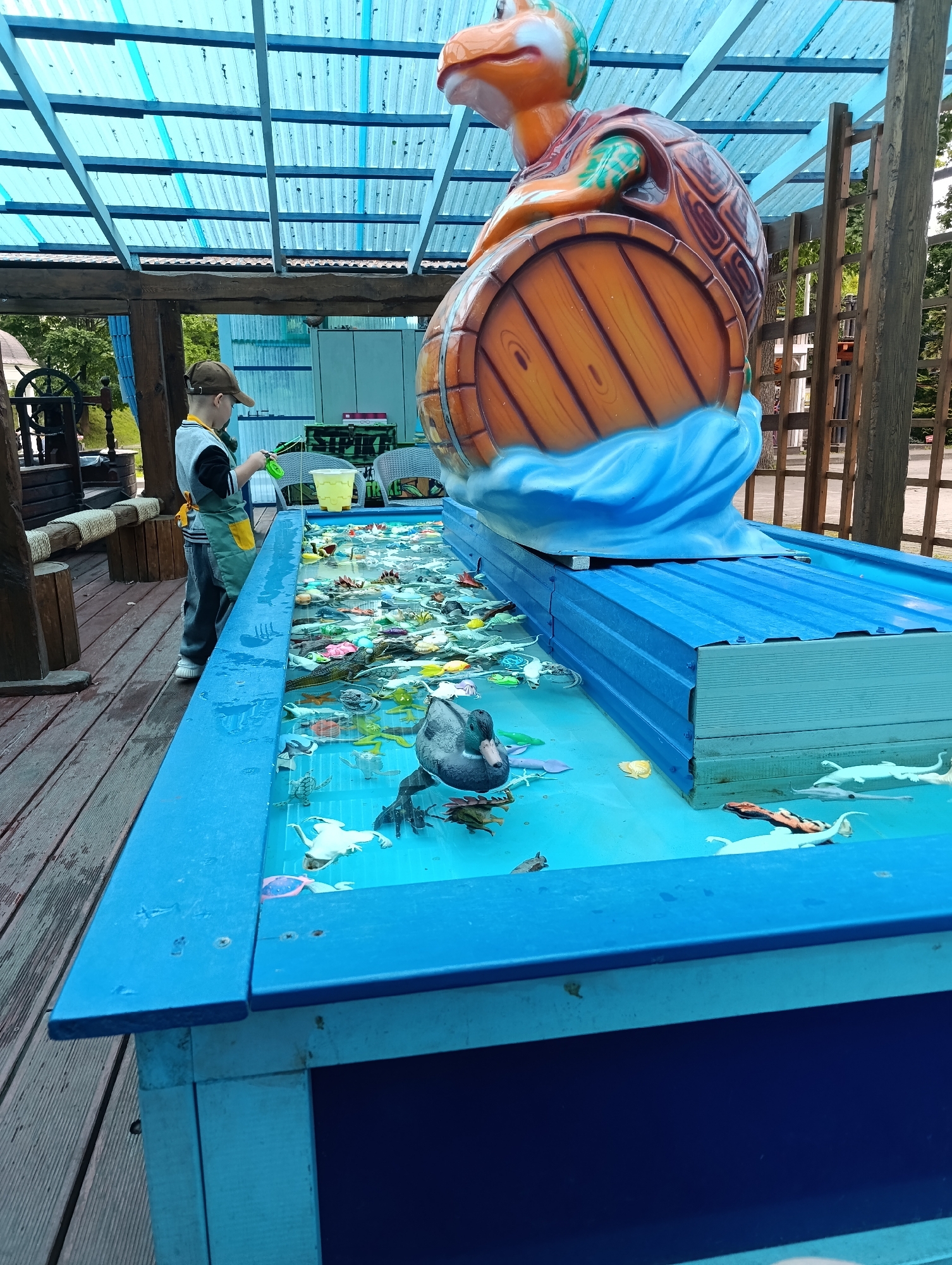 A boy wearing a cap and apron stands beside a blue pool filled with plastic sea creatures and toys. He is holding a green fishing rod, appearing to be engaged in a game. The pool is part of an outdoor attraction, possibly an amusement park or a public recreational area, with a large turtle sculpture mounted on a barrel in the background. The environment appears to be in Kaliningrad, Russia, given the context of previous images.
