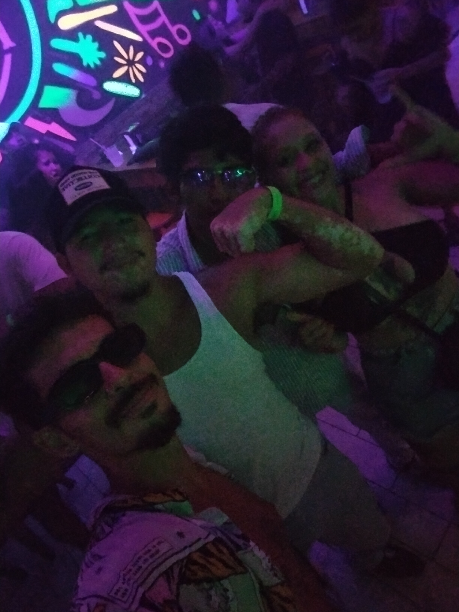 The image captures a group of four people in a brightly lit nightclub in Playa del Carmen, Mexico. The fluorescent lights highlight a mural on the back wall composed of musical notes and shapes. The individual nearest the camera is wearing sunglasses and a colorful shirt. To the rear is a man with a baseball cap and another man wearing sunglasses. A woman with a glowing green wristband is on the right, holding up a peace sign.