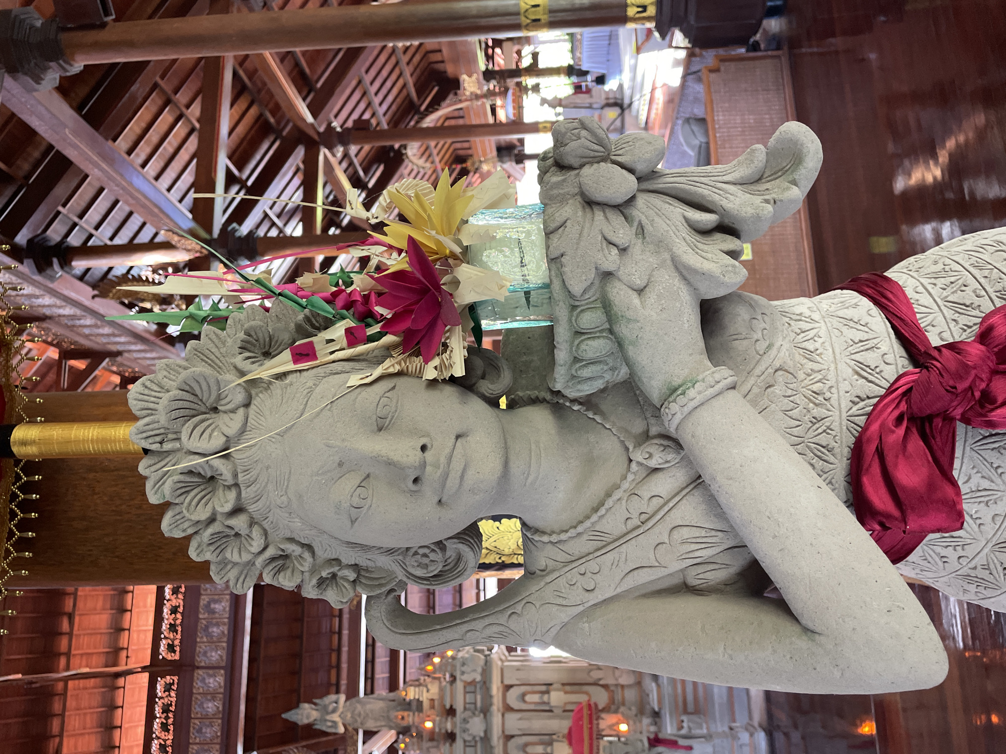 A stone statue of a woman wearing a floral headdress and a red sash is prominently displayed in the foreground. The statue holds a glass container filled with colorful, decorative paper flowers and palm fronds. Behind the statue, the interior of a building with a wooden beamed ceiling and decorative elements is visible. In the background, other statues and furniture are partially obscured by light and shadow. The overall scene suggests an indoor cultural or religious setting.